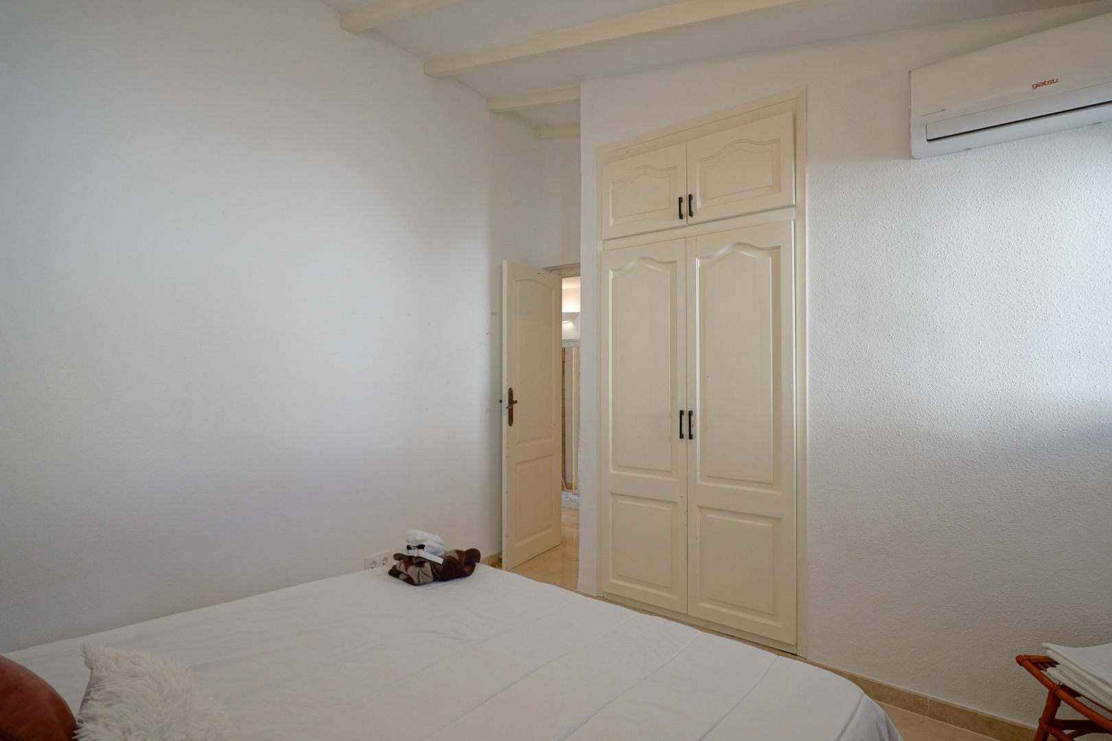 Helles Schlafzimmer mit Einbauschrank im klassischen Stil, sichtbaren Deckenbalken und einer wandmontierten Klimaanlage.