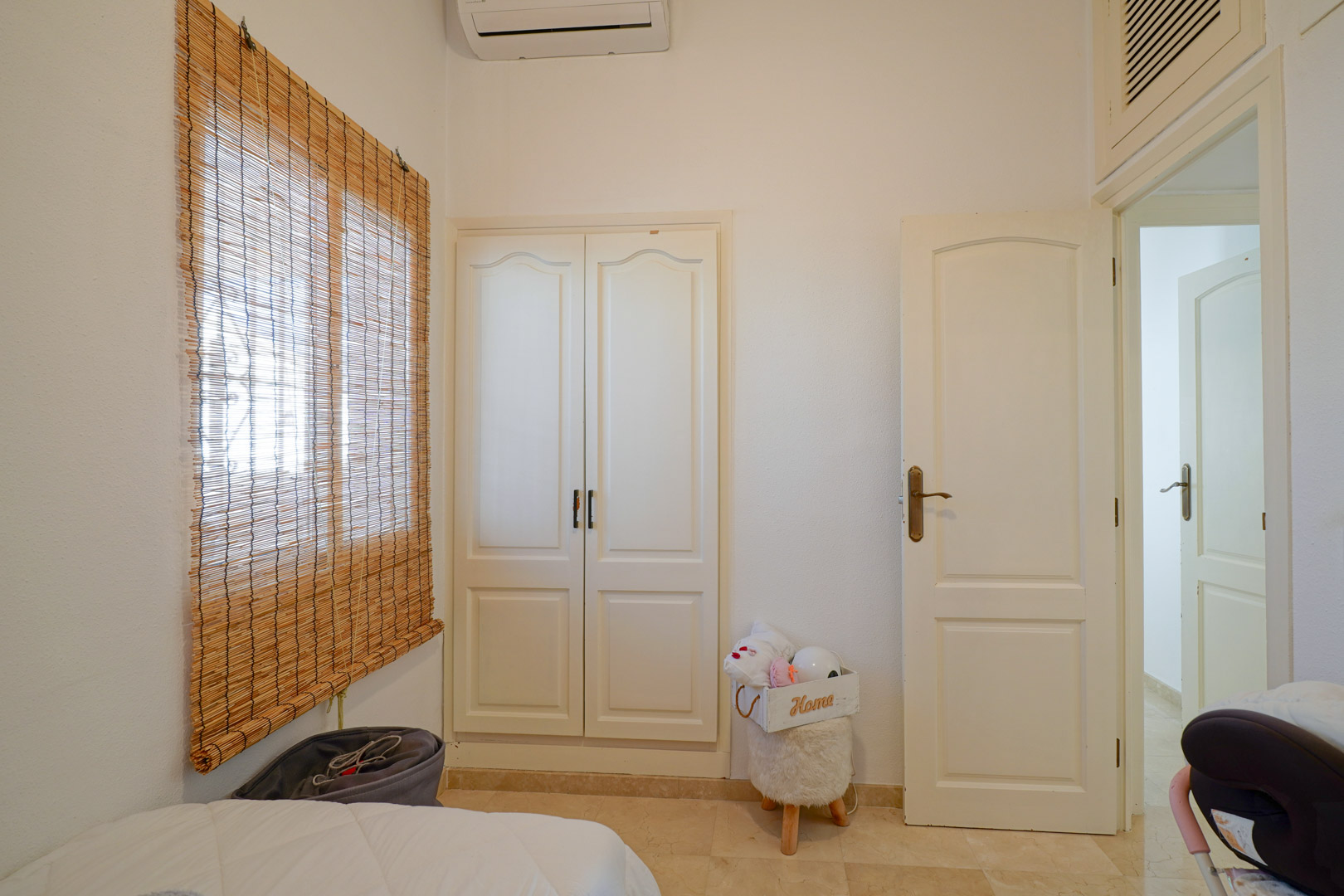 Helles Schlafzimmer mit weißem Einbauschrank, Split-Klimaanlage und Fenster mit Bambusrollo über hellem Fliesenboden.