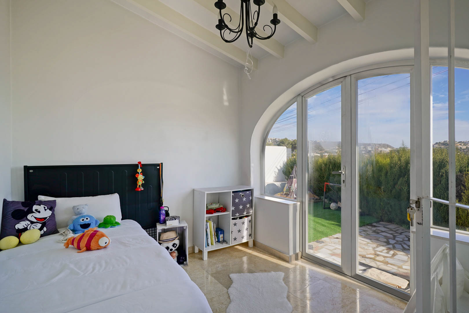 Helles Schlafzimmer mit weißen Balkendecken und einer großen bogenförmigen Glastür, die direkten Zugang zum Garten und viel Tageslicht bietet.