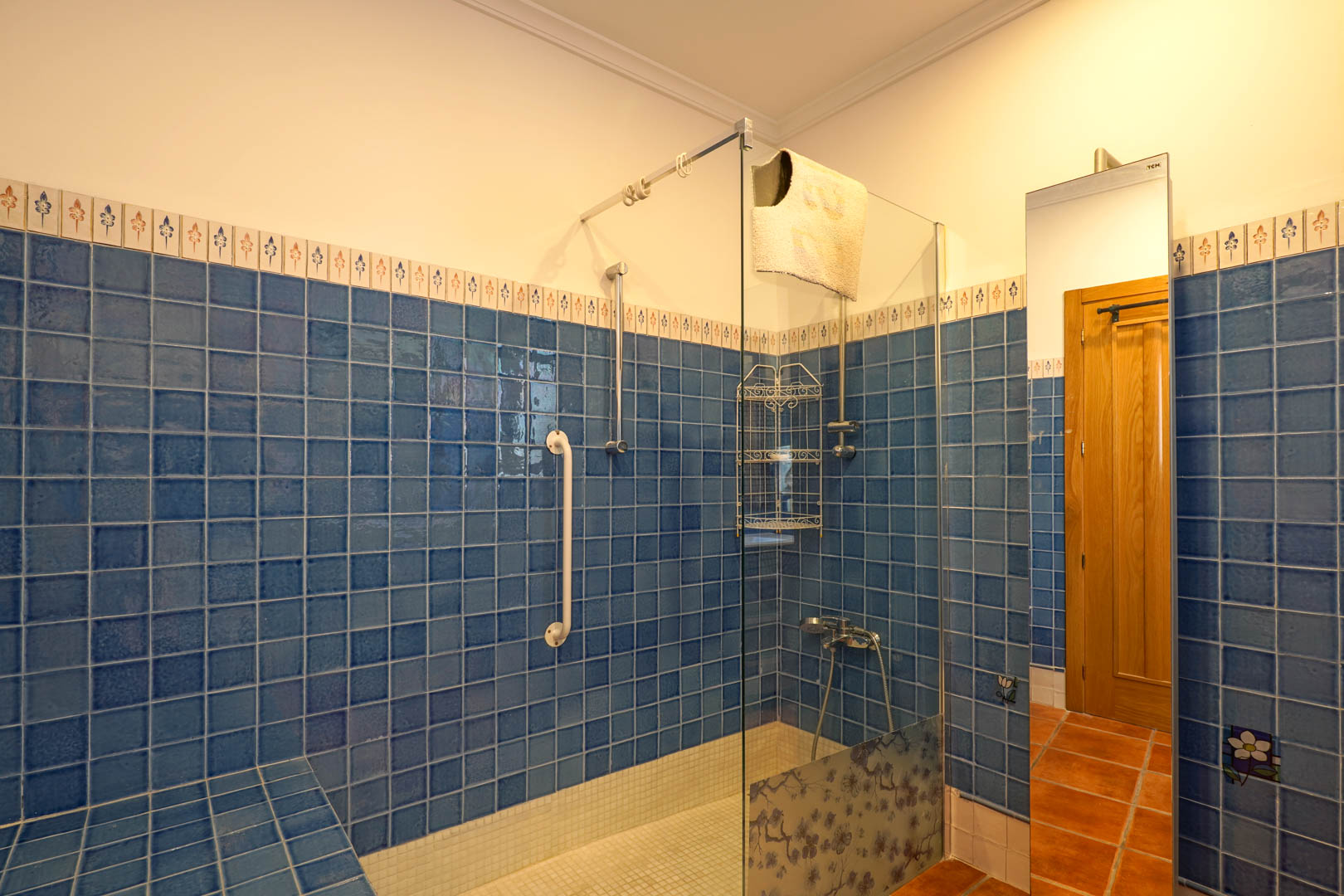Badezimmer mit begehbarer Dusche, blauen Fliesen, Glasabtrennung mit Blumenmuster, weißem Mosaikboden und einem Sicherheitsgriff an der Wand.