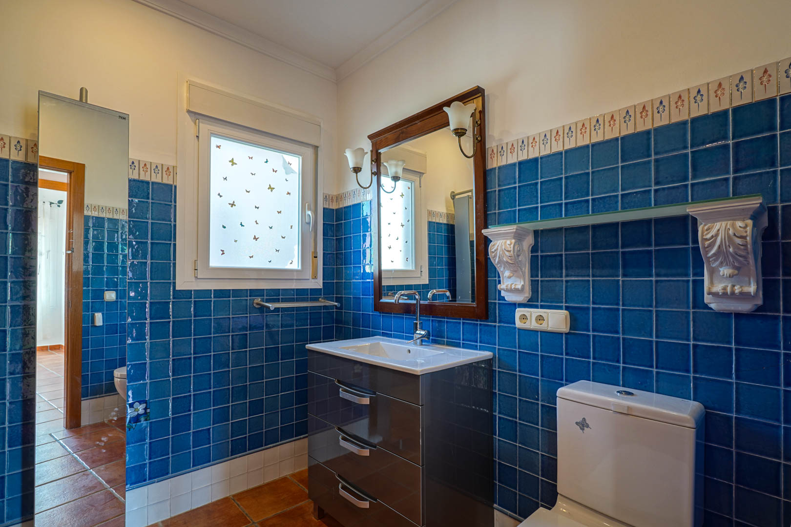 Badezimmer mit blauen Wandfliesen und dekorativer Bordüre, modernem Waschtisch, Holzrahmen-Spiegel und traditionellen Terrakotta-Bodenfliesen.