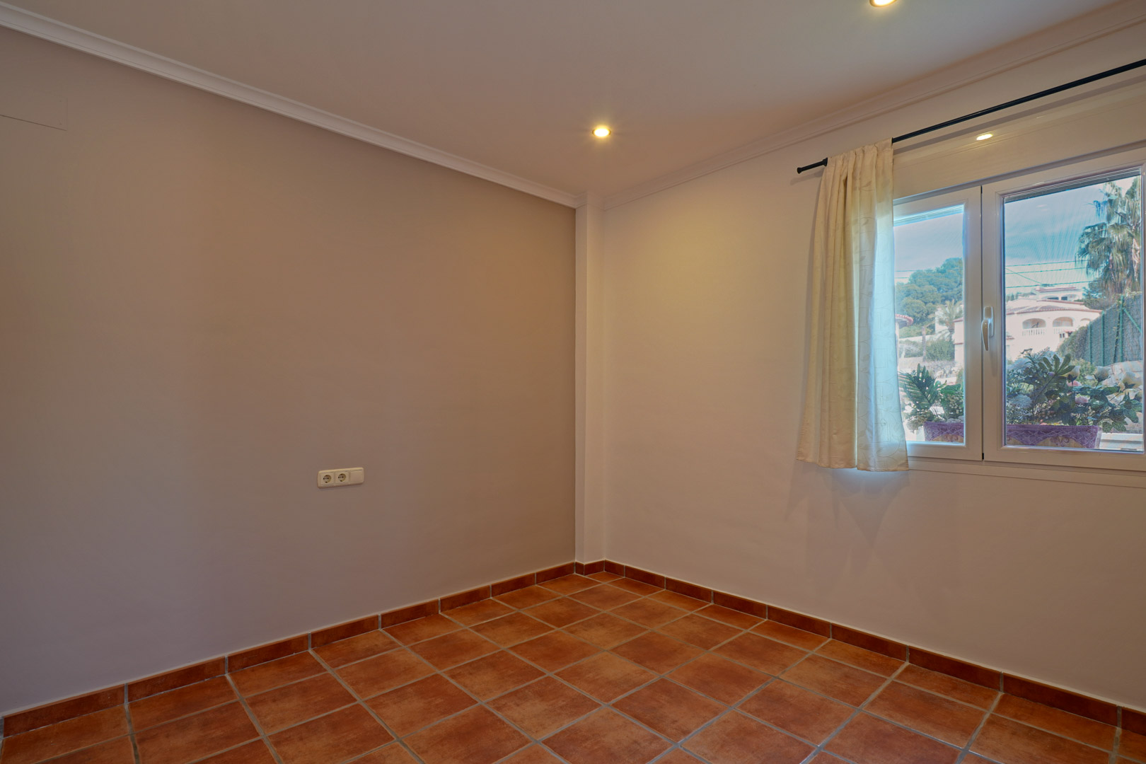 Helles Schlafzimmer mit rustikalen Terrakotta-Fliesen, neutralen Wänden und einem weißen PVC-Fenster mit Blick auf die mediterrane Umgebung.