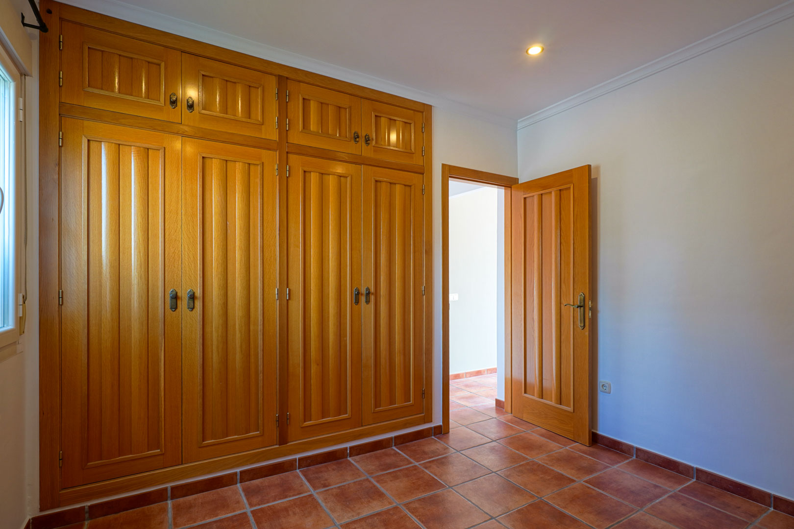 Schlafzimmer mit großem, deckenhohem Einbauschrank aus Holz, rustikalem Terrakotta-Fliesenboden und dezenter Deckenbeleuchtung.