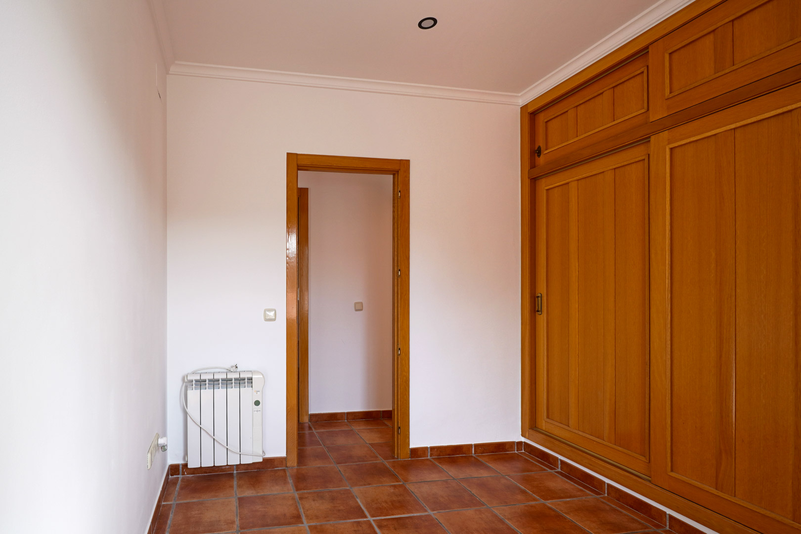 Schlafzimmer mit großem Einbauschrank aus Holz, rustikalen Terrakotta-Fliesen und Elektroheizkörper. Der Raum bietet weiße Wände, Deckenleisten und Zugang zum Flur.