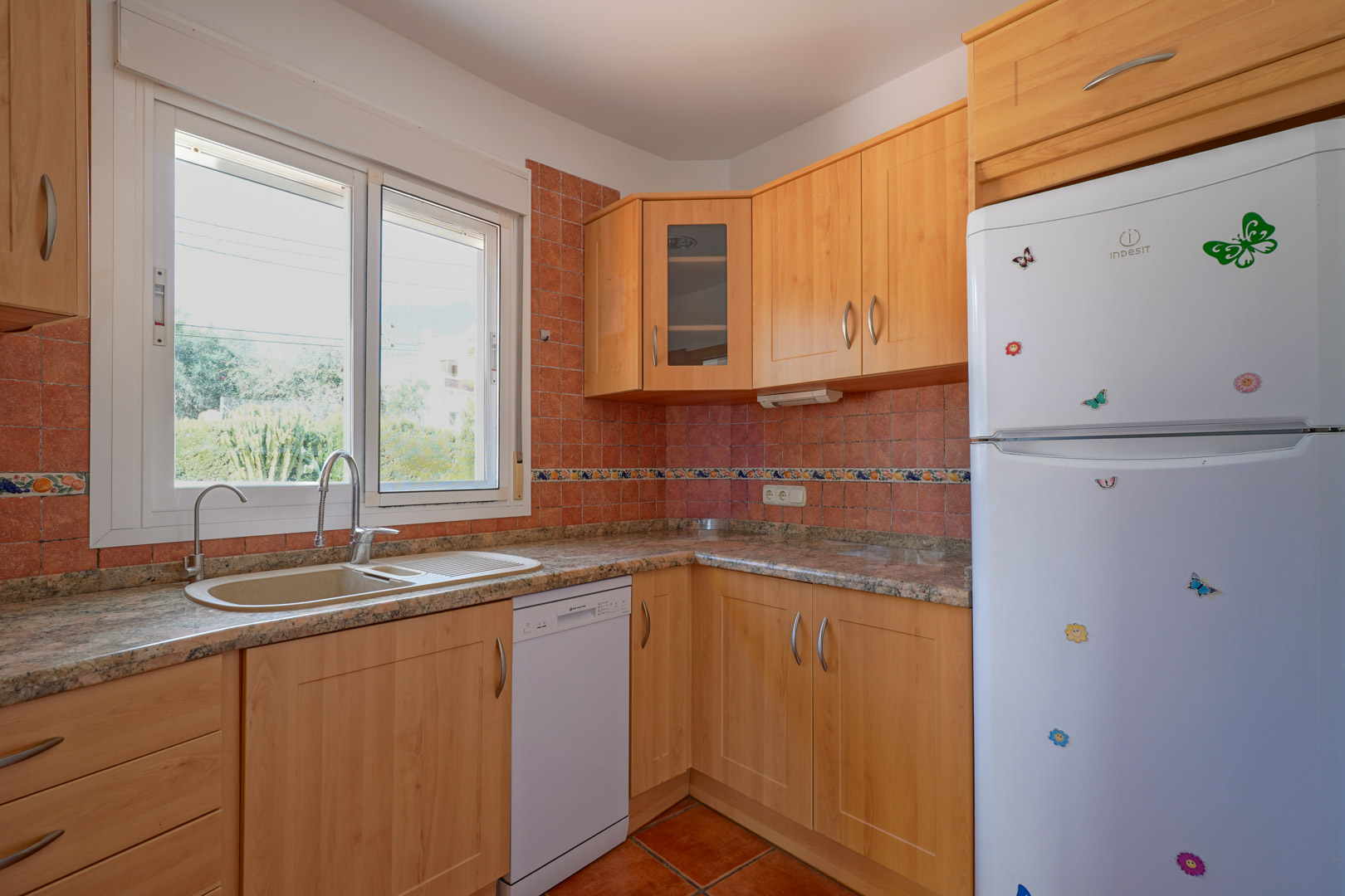Separate Küche mit hellen Holzschränken, Granitarbeitsplatte und rustikalen Fliesen. Ausgestattet mit Spülmaschine, Kühlschrank und großem Fenster für viel Tageslicht.