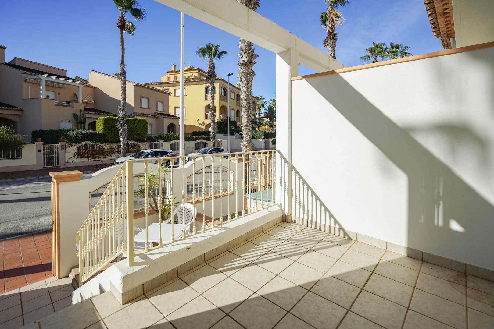 Erhöhte vordere Terrasse mit Keramikfliesen und Metallgeländer, mit Blick auf die palmengesäumte Straße und die umliegende Wohngegend.