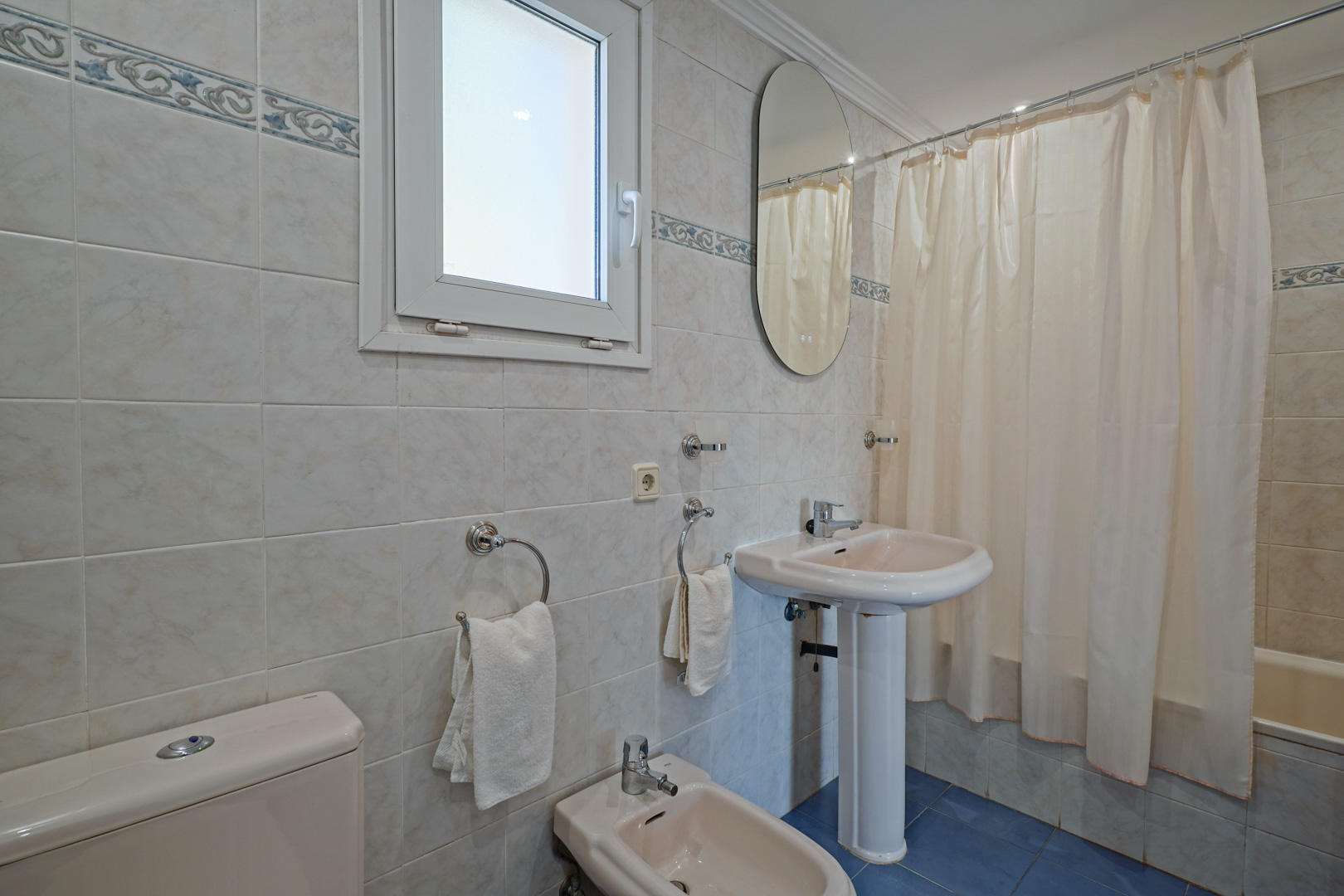 Familienbadezimmer mit cremefarbenen Sanitäranlagen, Standwaschbecken und Bidet. Ausgestattet mit einer Badewanne, blauen Bodenfliesen und einem Fenster für natürliches Licht.