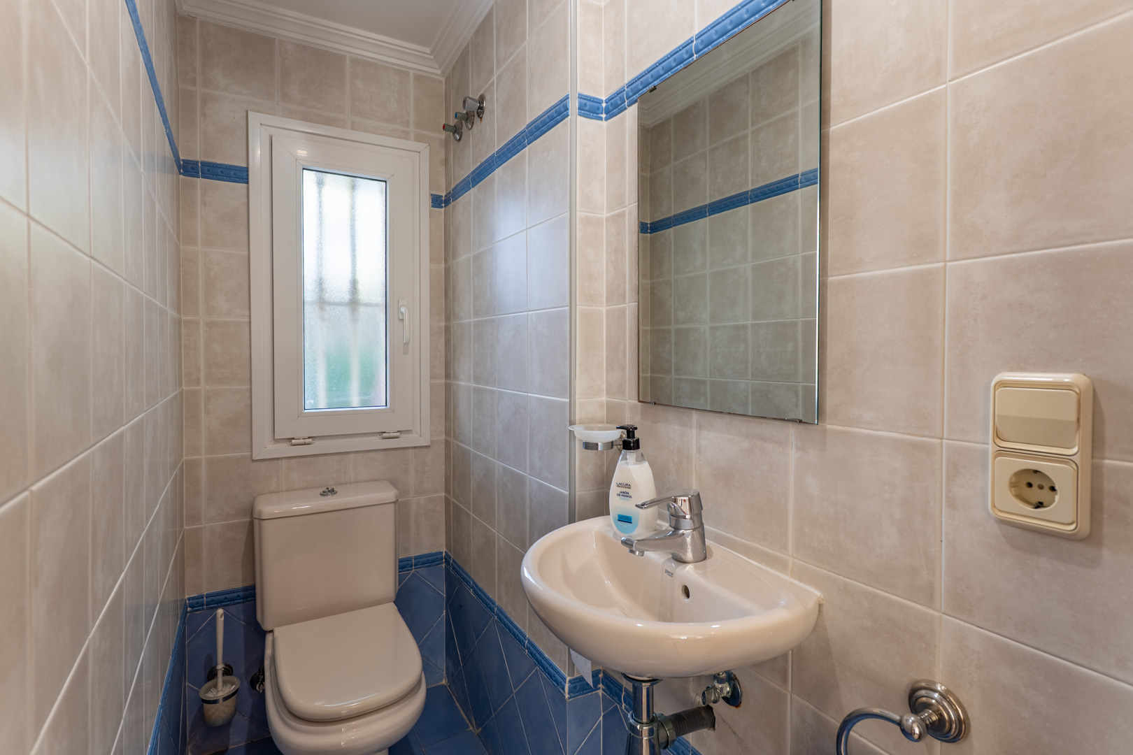 Gäste-WC mit beige-blauen Keramikfliesen, kompaktem Waschbecken und einem Fenster mit Milchglas für natürliches Licht und Privatsphäre.