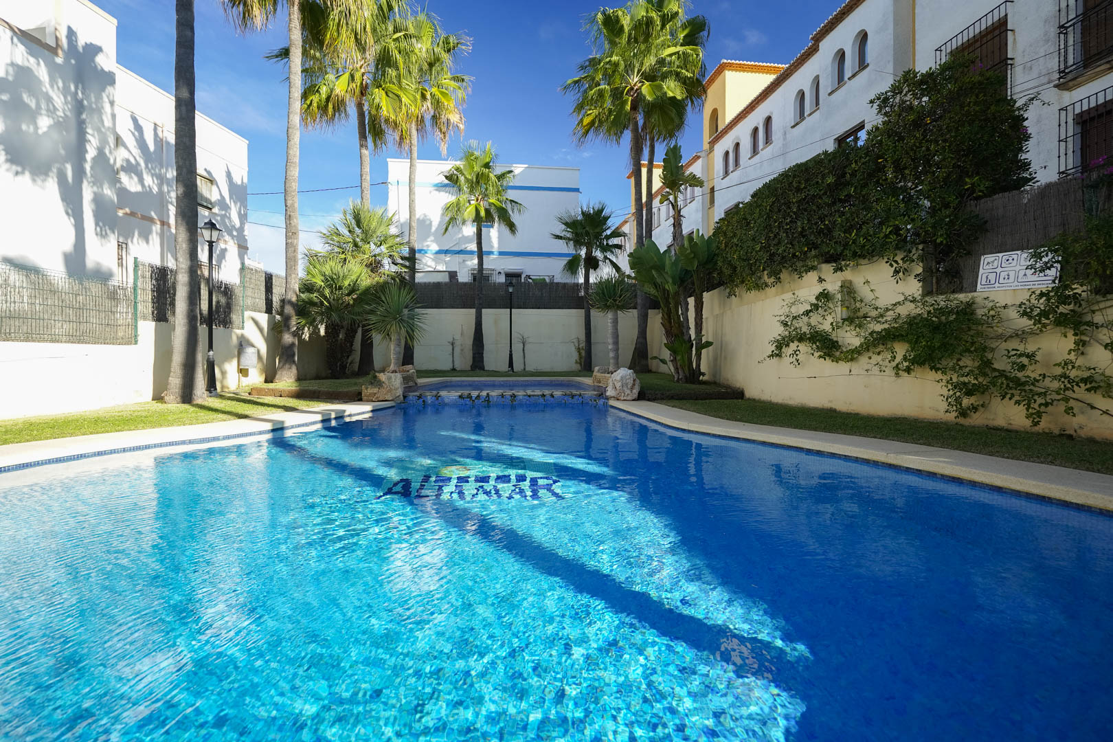 Gemeinschaftlicher Pool mit blauen Fliesen, umgeben von Palmen und Rasenflächen. Die Anlage bietet eine ruhige, sonnige Atmosphäre in einer gepflegten Wohnumgebung.