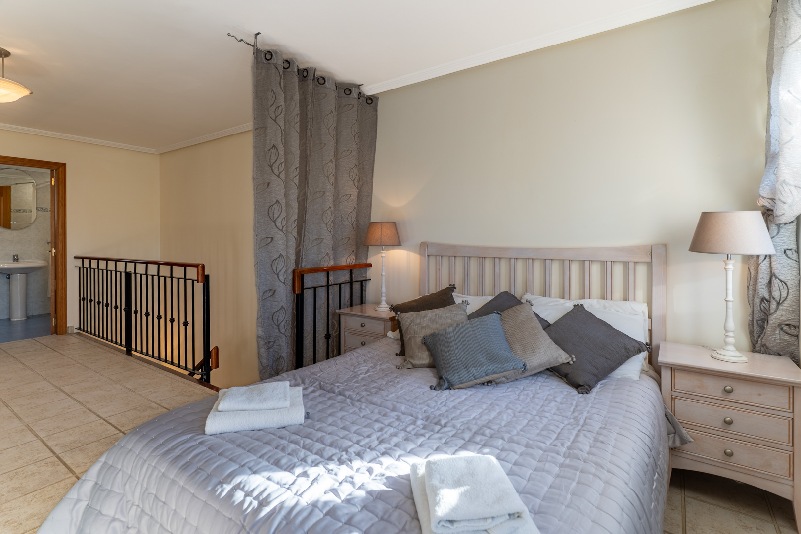 Hauptschlafzimmer im Obergeschoss mit En-Suite-Bad, Fliesenboden und einem dekorativen Metallgeländer am Treppenaufgang.