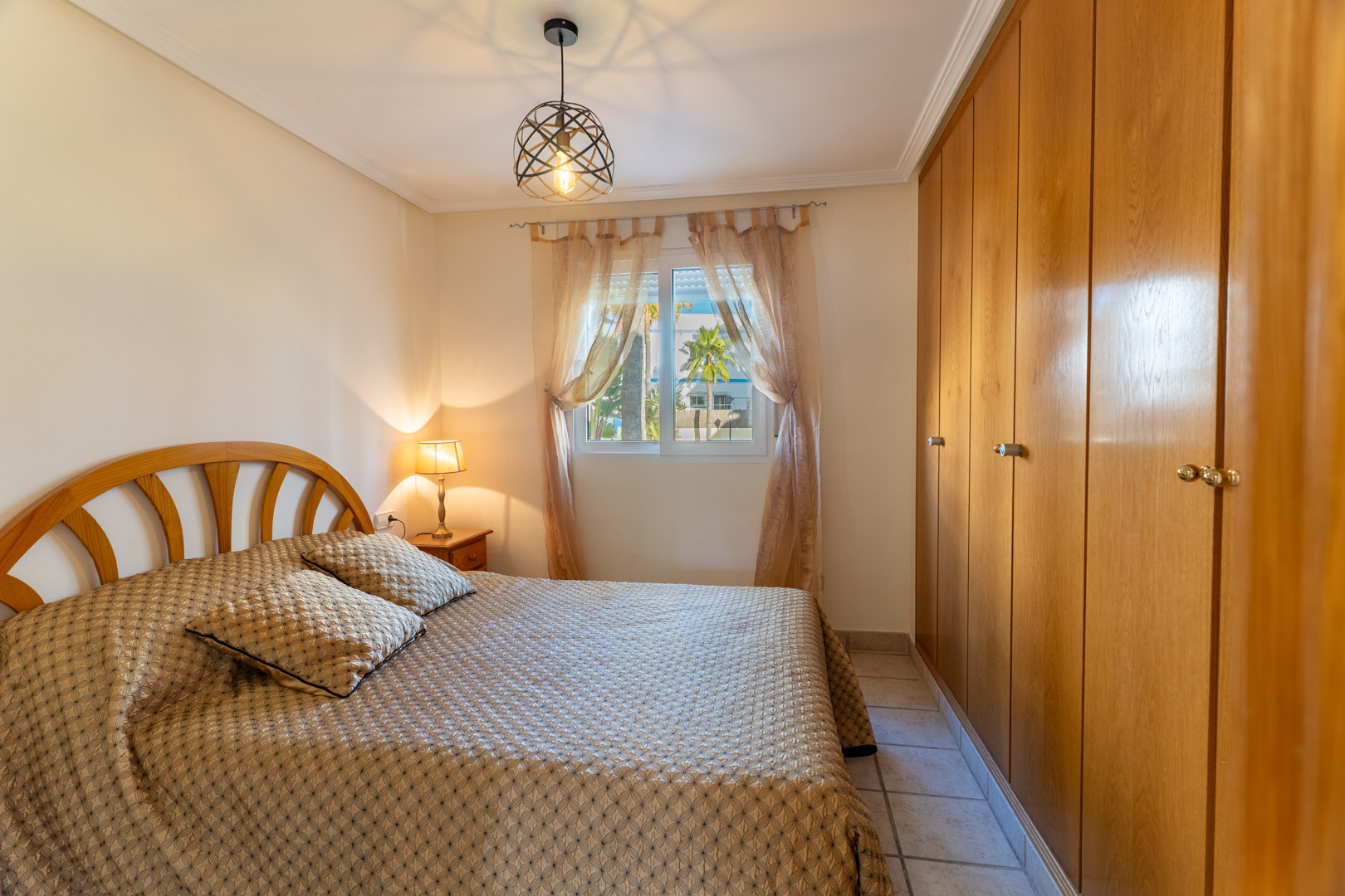 Helles Schlafzimmer mit großen Einbauschränken aus Holz, Doppelbett und Fenster mit Blick auf die Palmen der Wohnanlage.