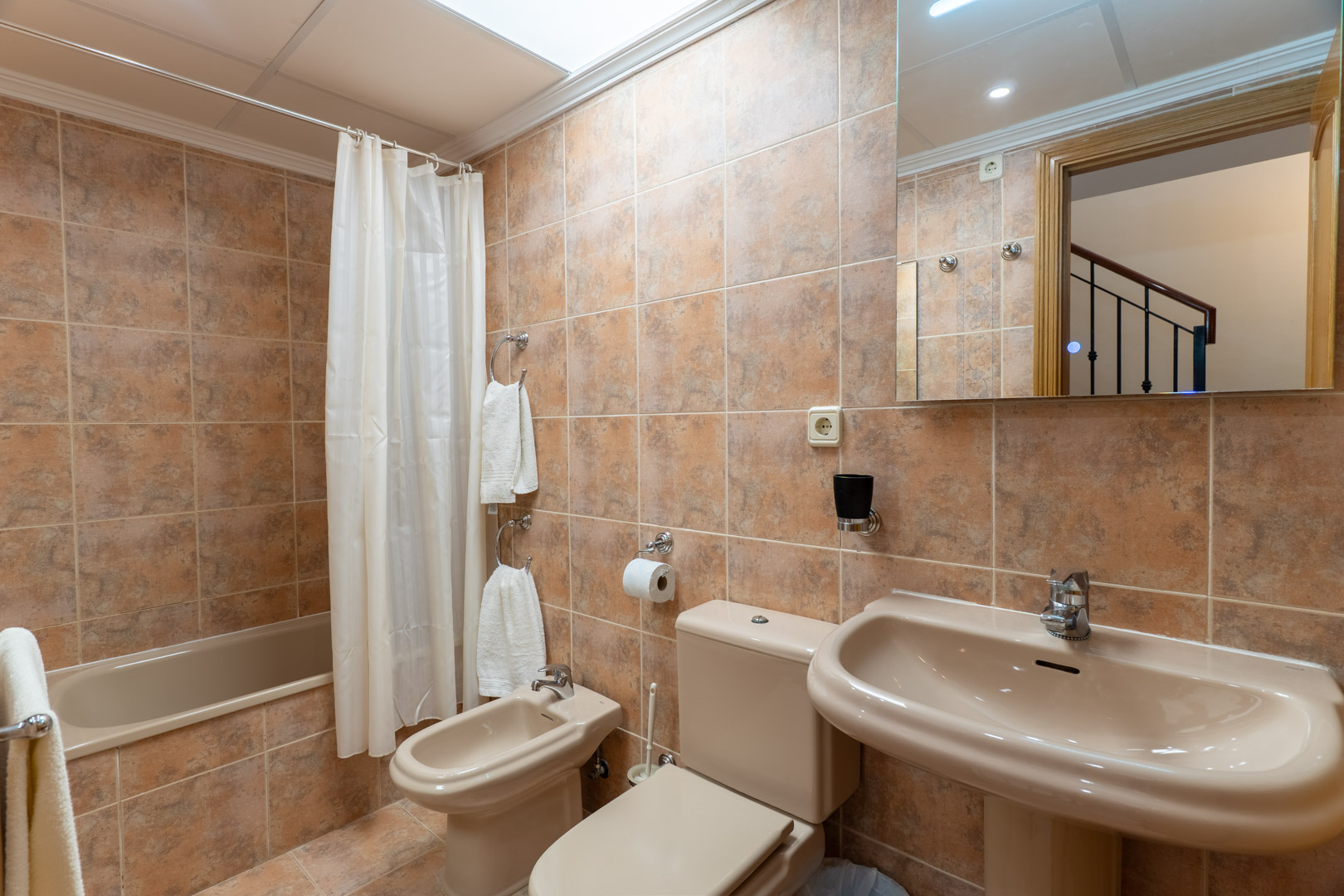 Komplettes Badezimmer mit Badewanne, Bidet, Standwaschbecken und erdfarbenen Keramikfliesen, beleuchtet durch ein Oberlicht.