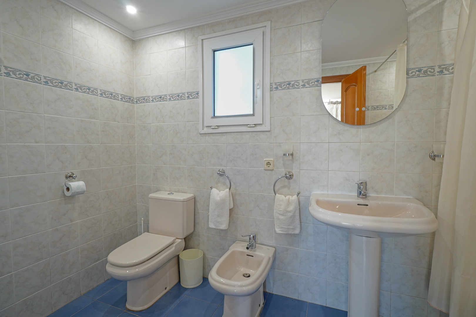 Komplettes Badezimmer mit hellen Wandfliesen, dekorativer Bordüre und blauem Boden. Ausgestattet mit WC, Bidet, Standwaschbecken und einem Fenster.