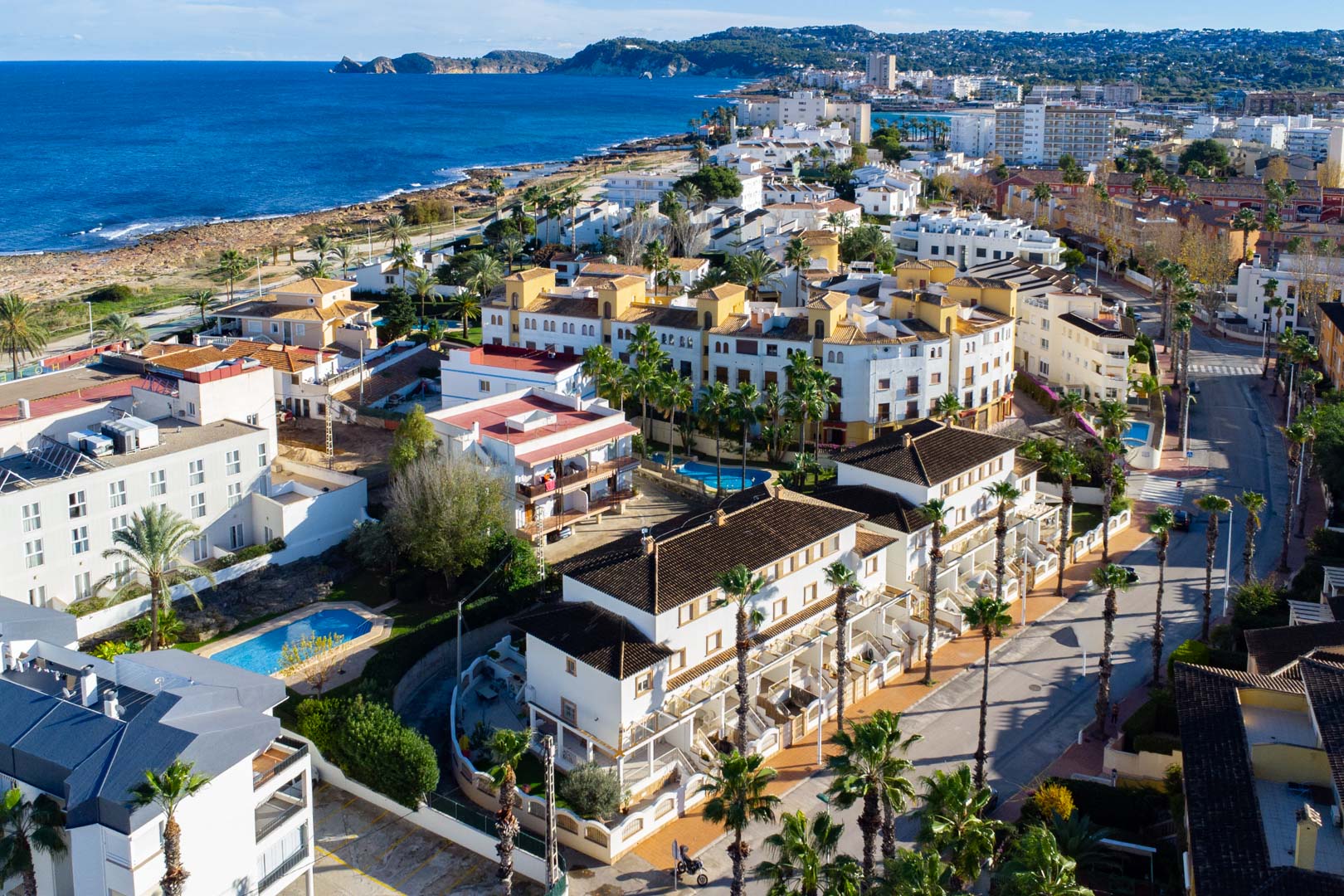 Luftaufnahme der Anlage in der Calle Génova, die die Nähe zum Mittelmeer, den Gemeinschaftspool und die Architektur mit weißen Fassaden und Palmen verdeutlicht.