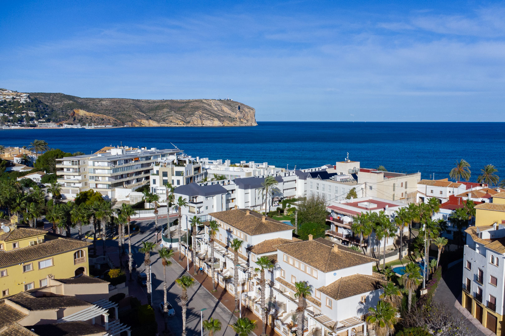 Panorama-Luftaufnahme von Jávea mit Blick auf das Mittelmeer und das Cabo de San Antonio, geprägt von traditionellen Ziegeldächern und von Palmen gesäumten Straßen.