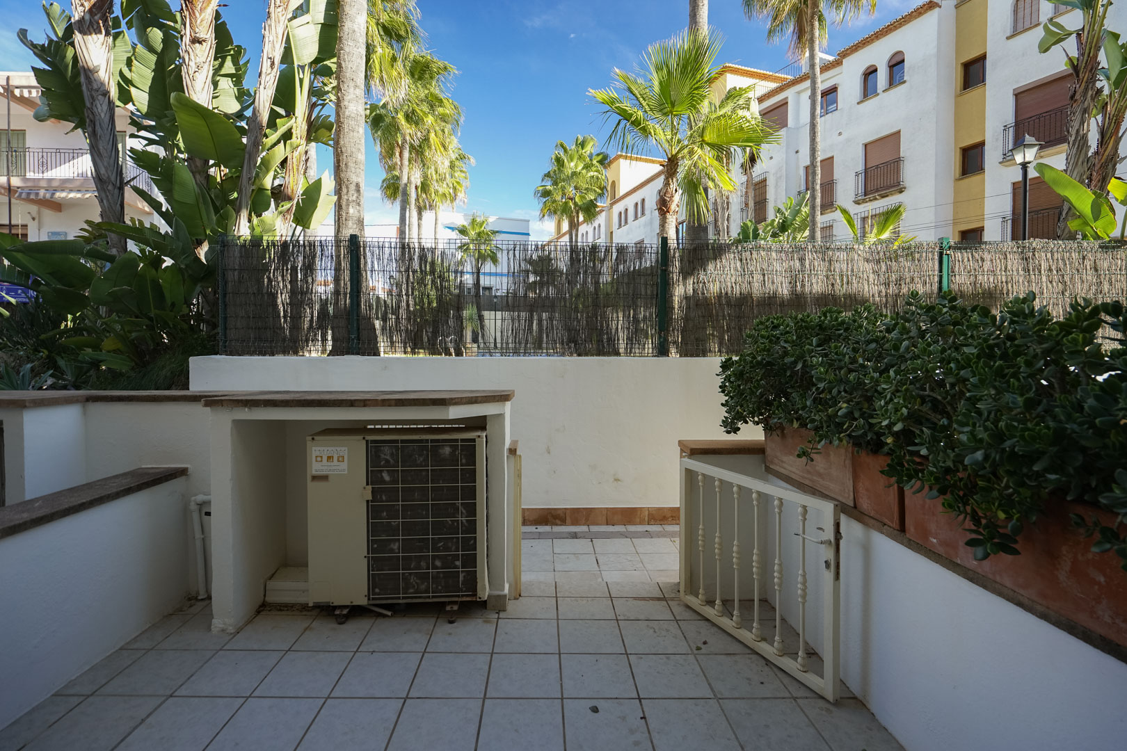 Private geflieste Terrasse mit integriertem Sichtschutz für die Klimaanlage, Pflanzkübeln und Blick auf die umliegenden Palmen und mediterrane Pflanzen.