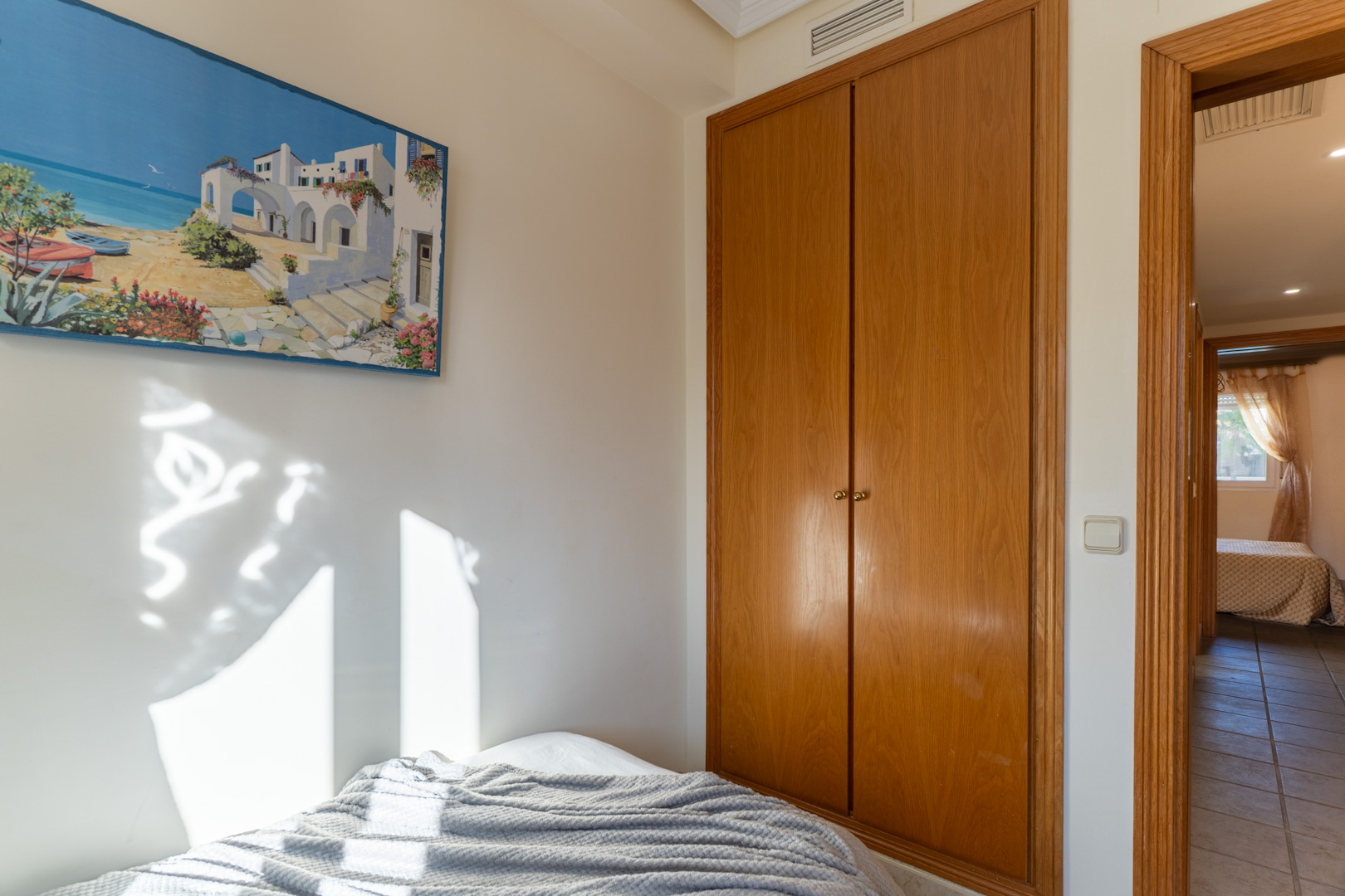 Schlafzimmer mit Einbauschrank aus Holz und natürlichem Lichteinfall. Der Türrahmen führt zu einem gefliesten Flur und einem weiteren Wohnbereich.