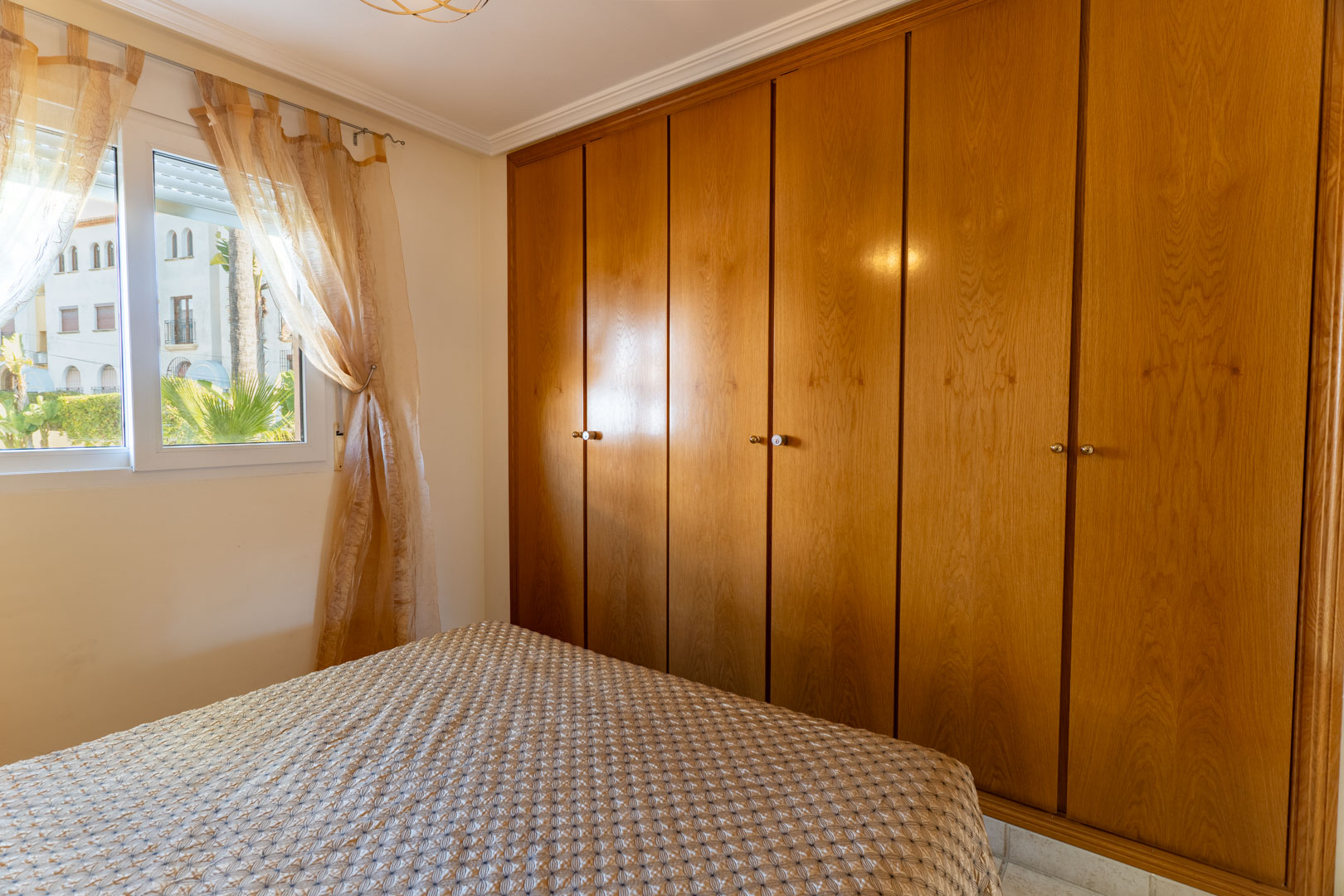 Schlafzimmer mit einem großen sechs-türigen Einbauschrank aus Holz und einem Fenster mit Blick auf die gepflegte Außenanlage und Tageslicht.