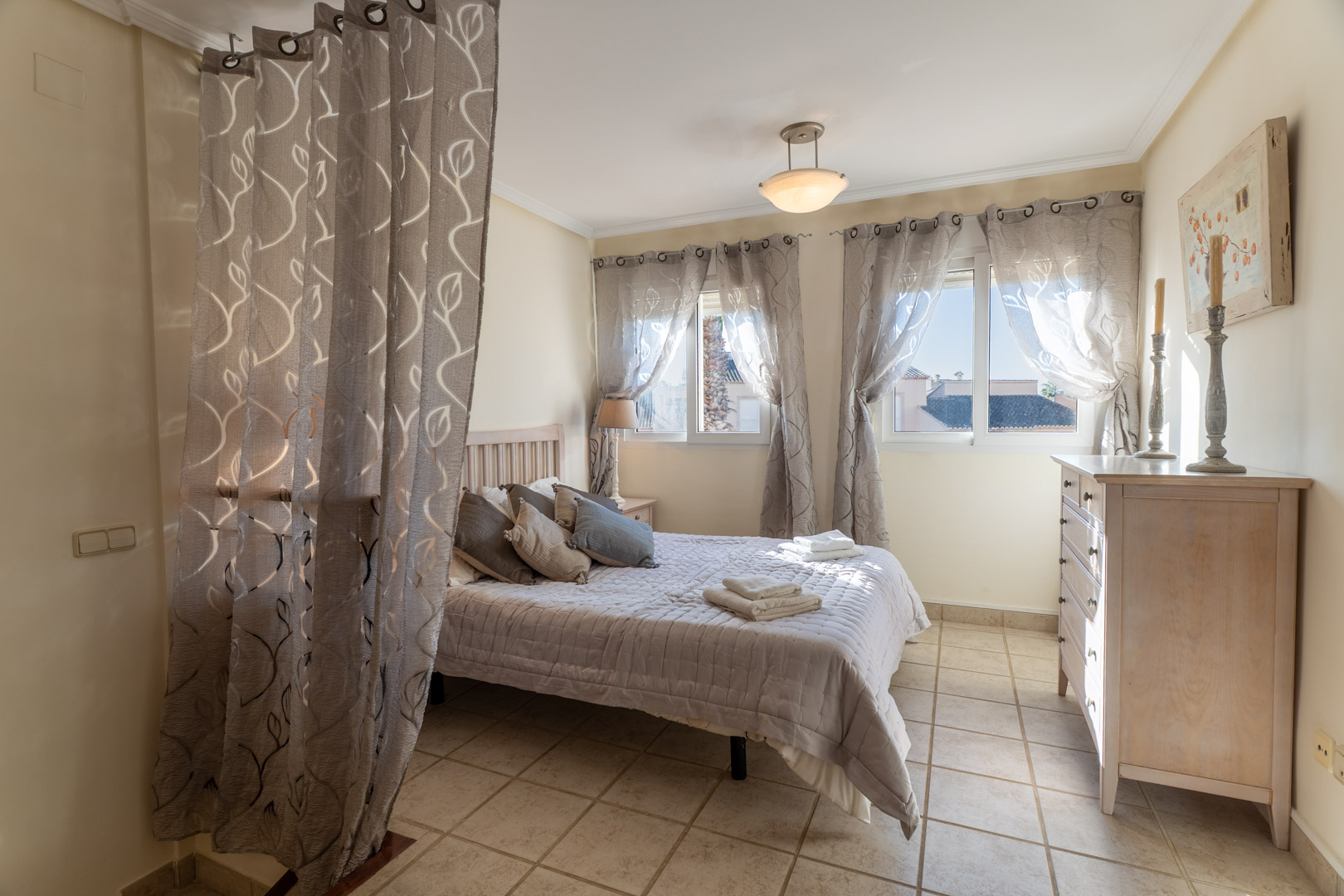 Schlafzimmer mit Fliesenboden, Doppelbett, Holzkommode und Fenstern mit gemusterten Vorhängen, die für natürliches Licht sorgen.