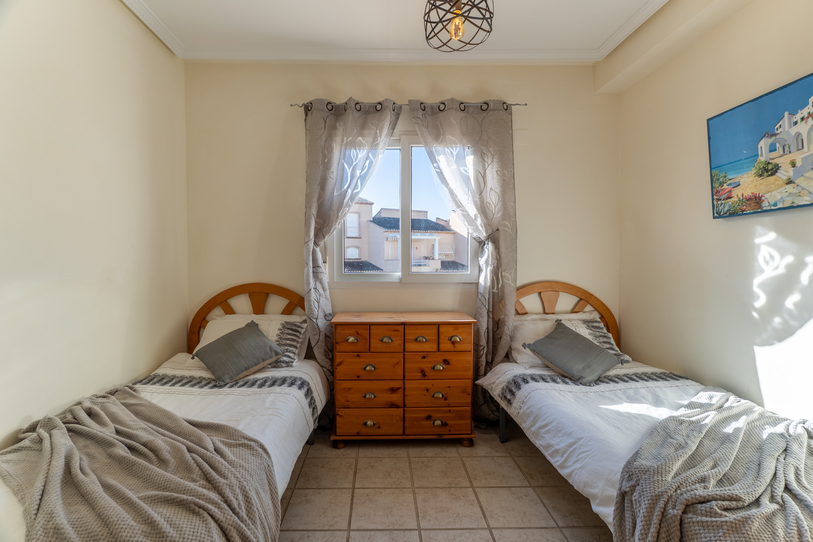 Zweibettzimmer mit hölzernen Kopfteilen und einer zentralen Kiefernkommode. Der Raum bietet Fliesenboden, natürliches Licht und eine moderne Drahtgitterleuchte.