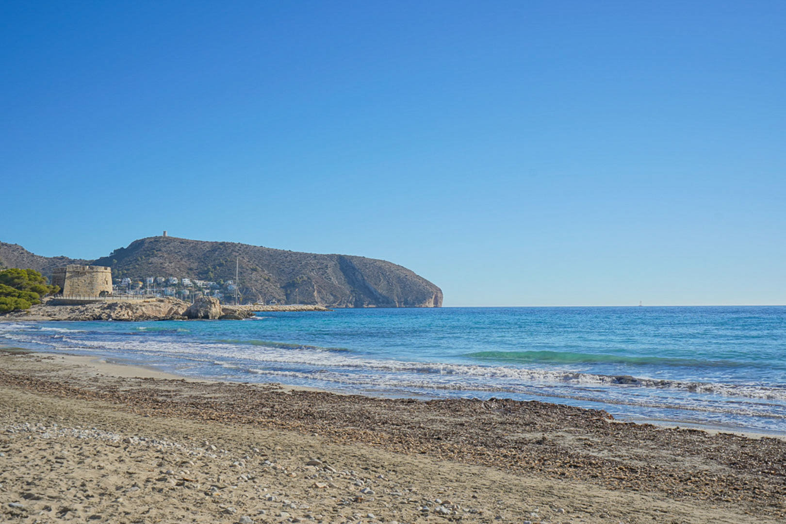 Blick auf den Strand von Moraira mit seinem historischen Turm und dem Mittelmeer unter klarem, blauem Himmel.