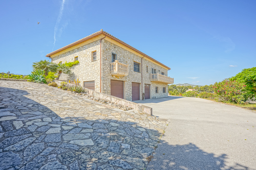 Außenansicht einer Villa mit Natursteinfassade, Dreifachgarage, gepflasterter Auffahrt und klassischen Steinbalkonen in mediterraner Umgebung.