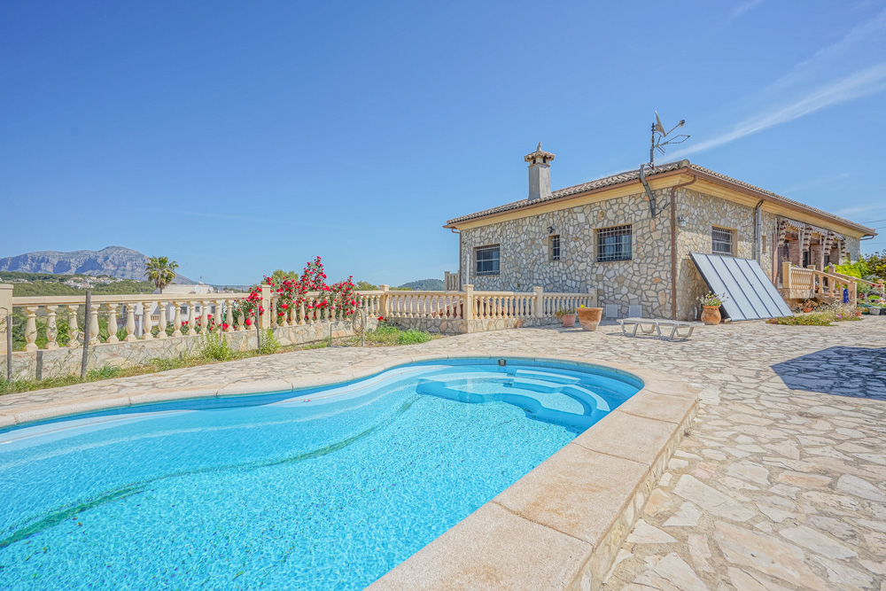Außenansicht mit geschwungenem Pool und einer Natursteinvilla. Die Terrasse mit Steinplatten bietet einen weiten Blick auf die Berge und die Landschaft.