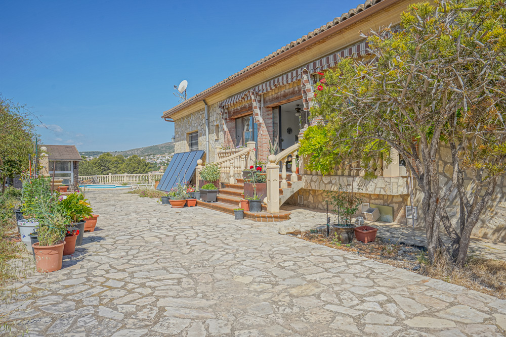 Außenansicht mit Natursteinpflaster, Steinfassade und Solarpanelen. Eine Treppe mit Balustrade führt zur überdachten Terrasse mit gestreiften Markisen.