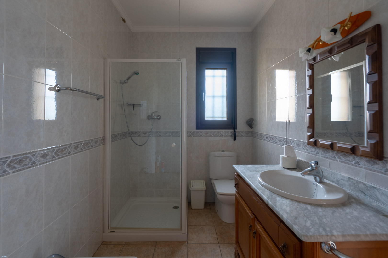 Badezimmer mit Marmorwaschtisch, Holzunterschrank und Glasduschkabine. Geflieste Wände mit dekorativer Bordüre und ein Fenster mit Sichtschutzglas.