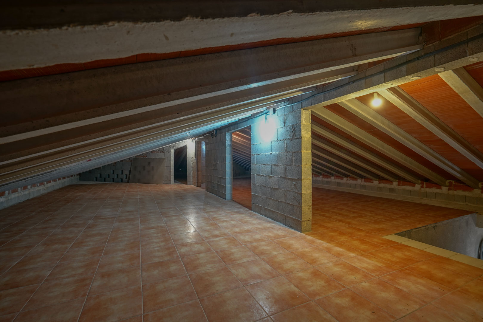 Dachboden-Stauraum mit terrakottafarbenen Bodenfliesen, sichtbaren Betondeckenbalken und Trennwänden aus Betonsteinen.