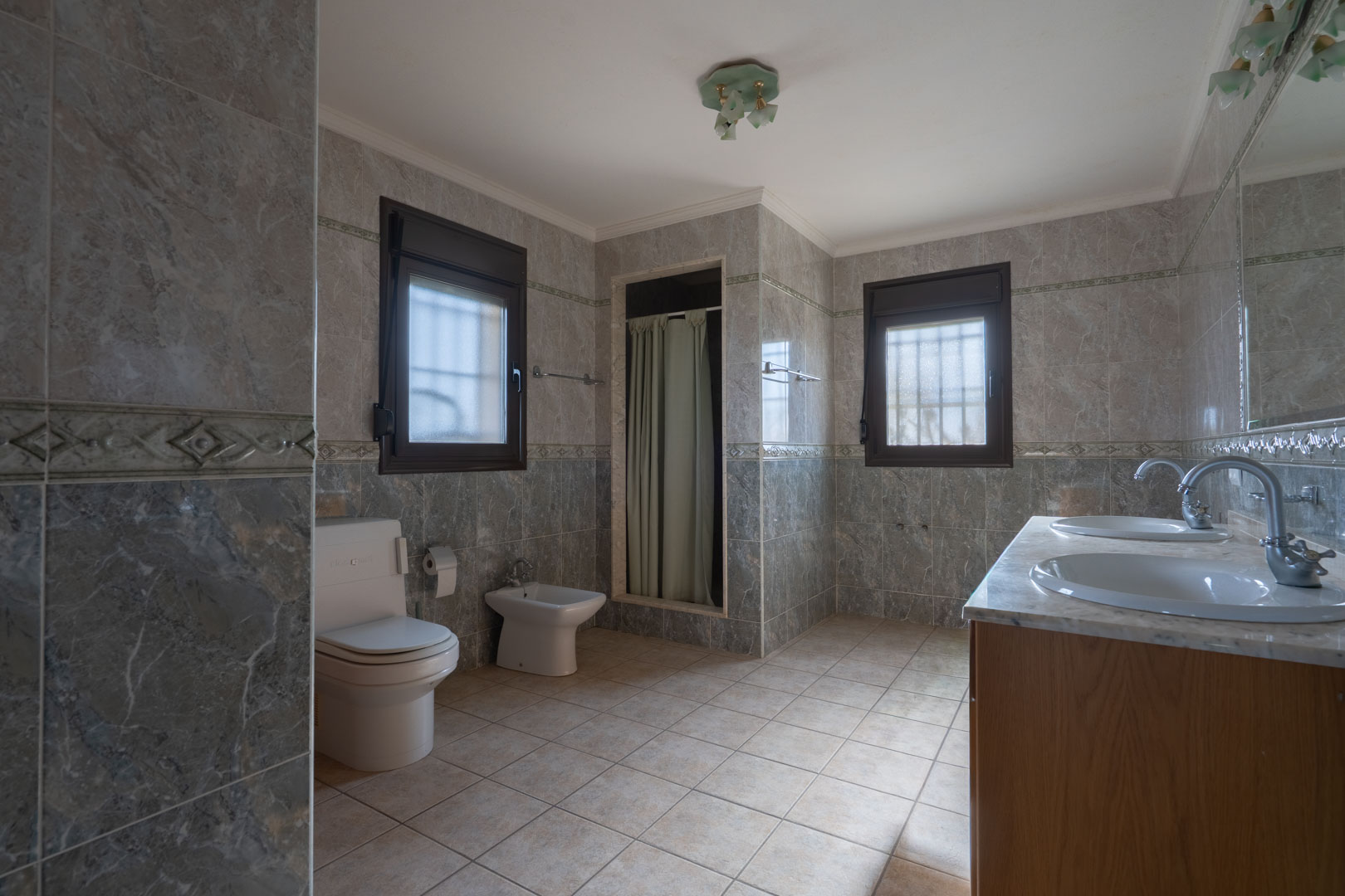 Geräumiges Badezimmer mit Doppelwaschtisch aus Marmor, grauen Wandfliesen mit Bordüre und zwei Fenstern mit dunklen Rahmen.