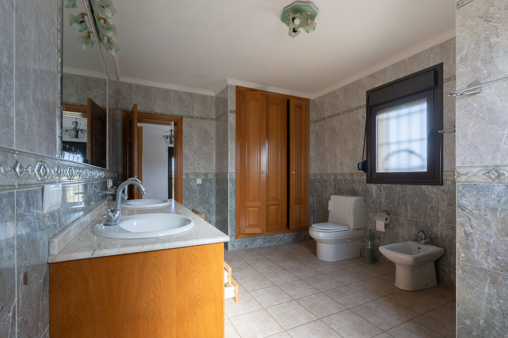 Großzügiges Badezimmer mit Doppelwaschtisch aus Marmor, Einbauschränken aus Holz und wandhohen Fliesen mit dekorativen Bordüren. Inklusive WC und Bidet.