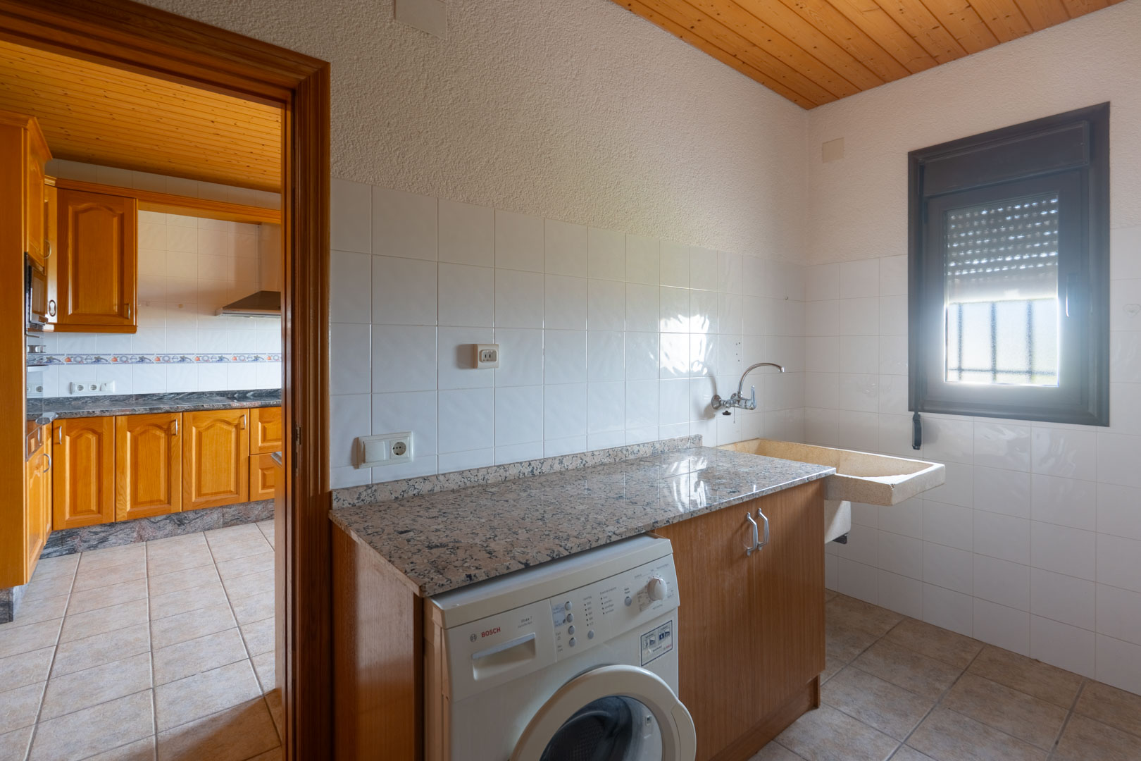 Hauswirtschaftsraum mit Granitarbeitsplatte, Bosch-Waschmaschine und Steinwaschbecken. Ausgestattet mit Holzdecke und weißen Fliesen, angrenzend an die Küche.