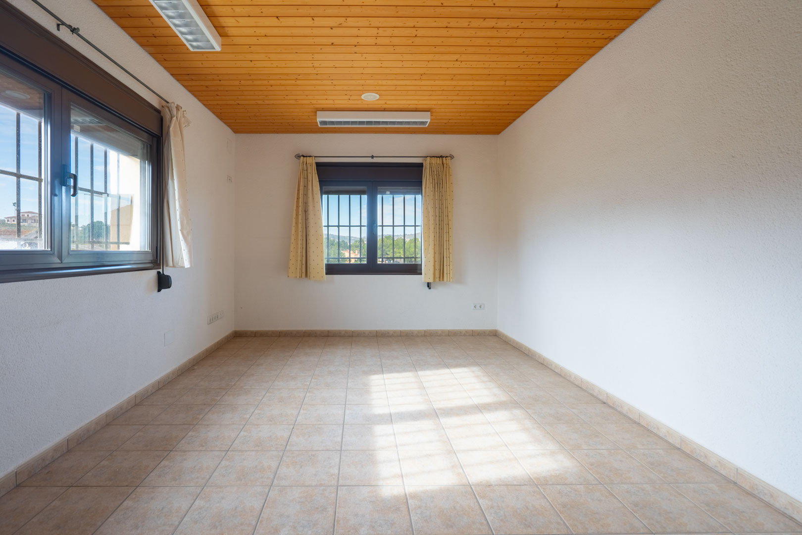 Heller, leerer Raum mit Holzdecke, beigem Fliesenboden und zwei Fenstern mit Sicherheitsgittern und cremefarbenen Vorhängen.