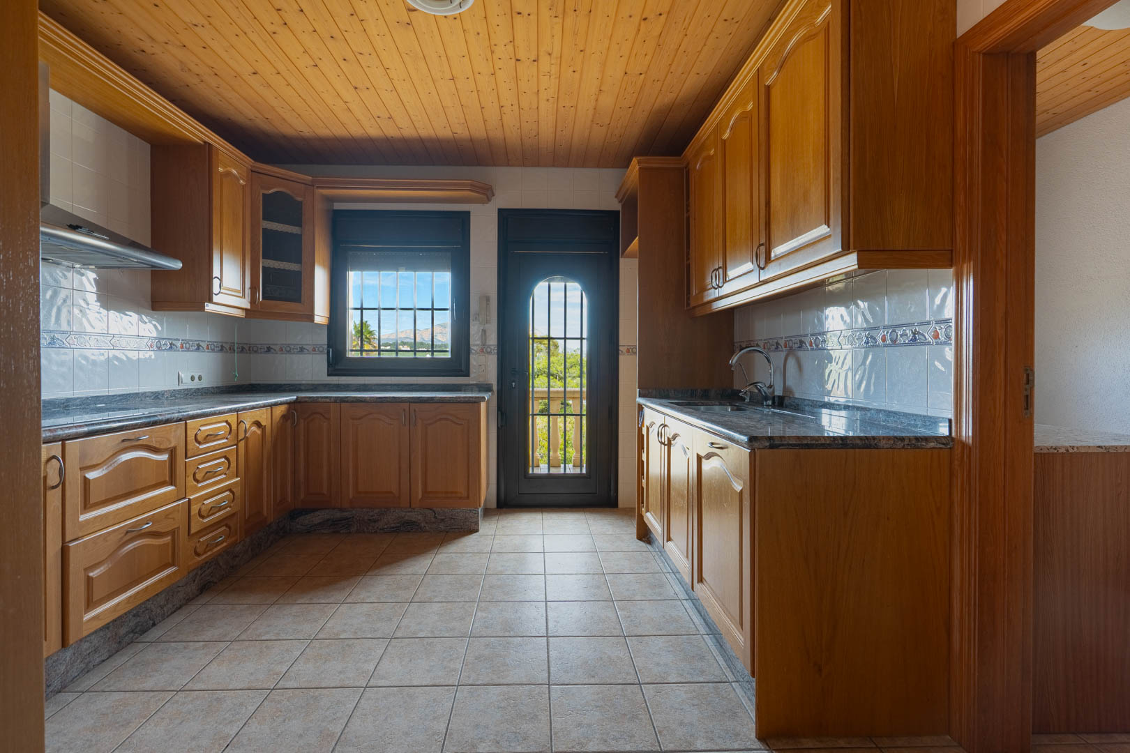 Küche mit Holzschränken, Granitarbeitsplatten und einer Holzdecke. Der Raum bietet Fliesenboden sowie Zugang zu einem Balkon mit Blick auf die umliegende Landschaft.