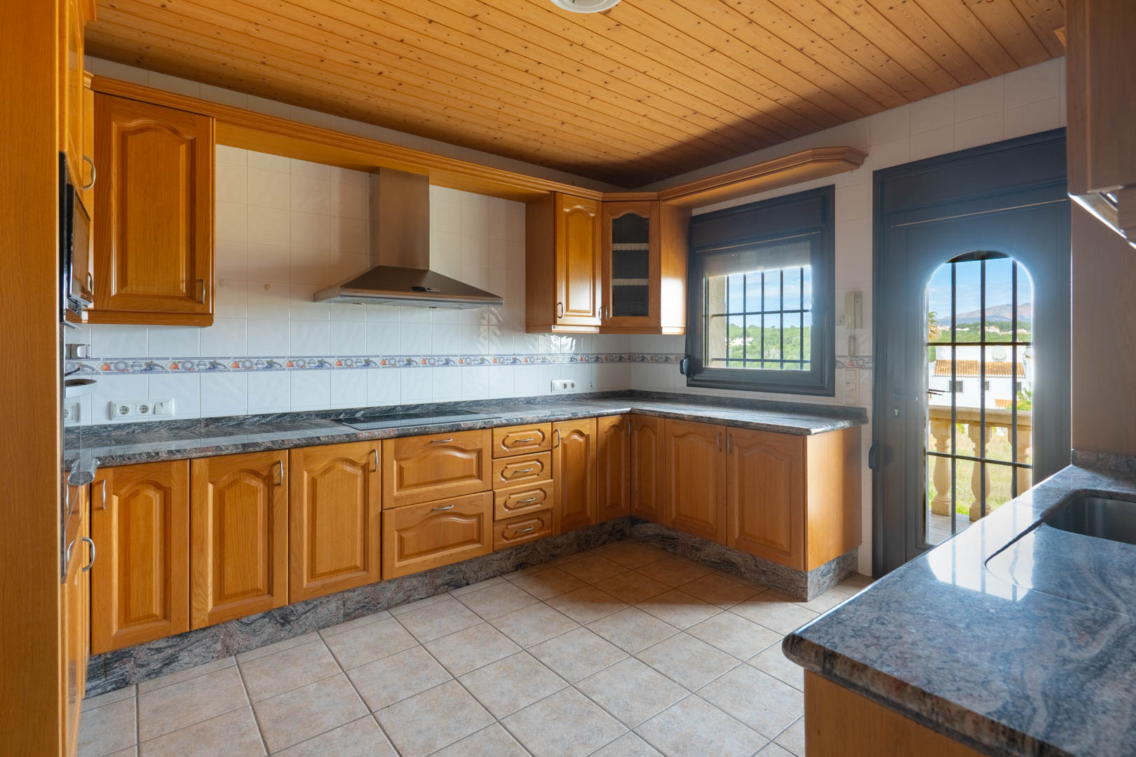 Küche mit Holzschränken, Granitarbeitsplatten und Holzdecke. Ausgestattet mit Edelstahlabzugshaube und Zugang zu einer Terrasse mit Blick auf die Landschaft.