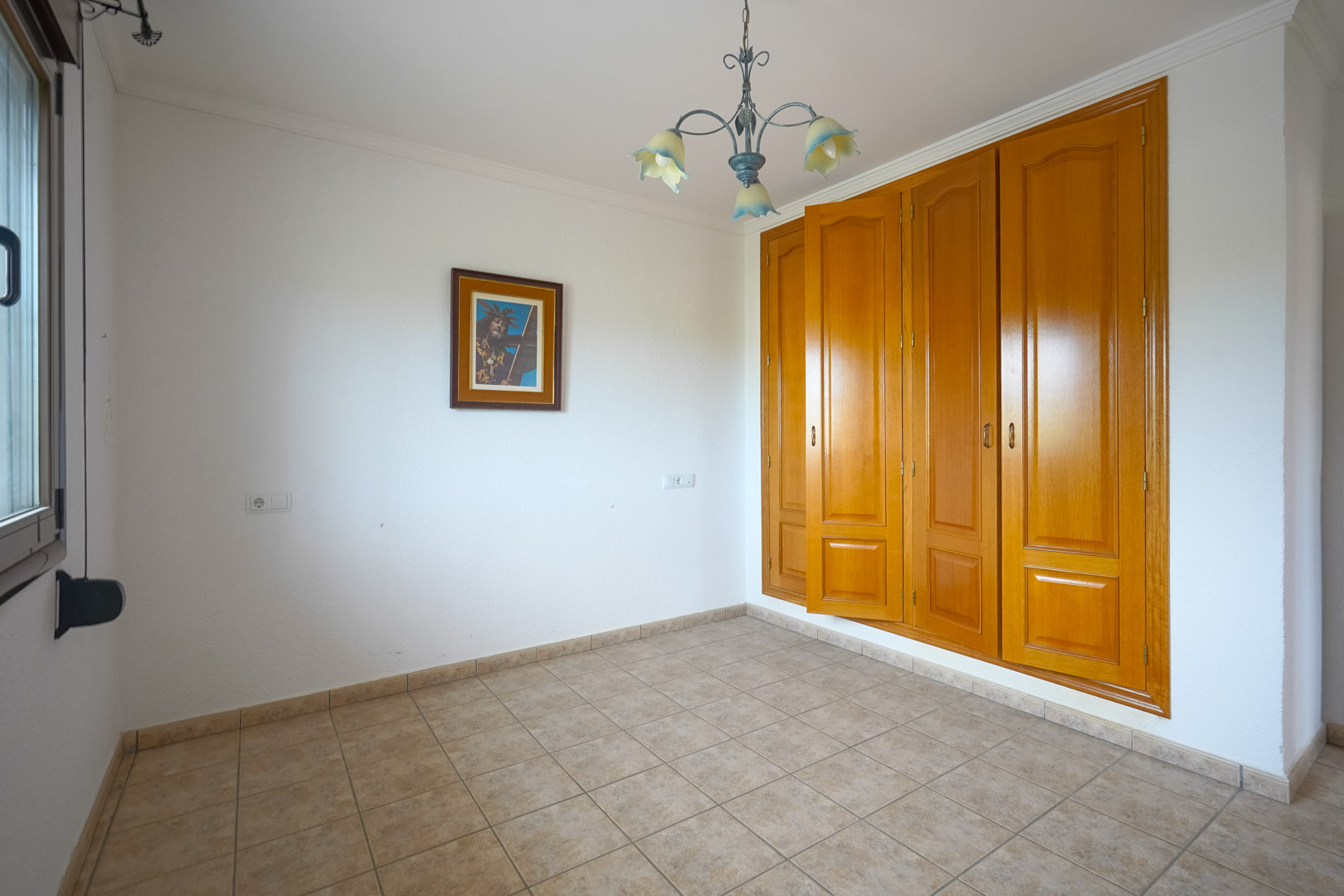 Leeres Zimmer mit einem großen viertürigen Einbauschrank aus Holz, beigen Keramikfliesen und einem dekorativen Deckenleuchter mit floralen Glasschirmen.