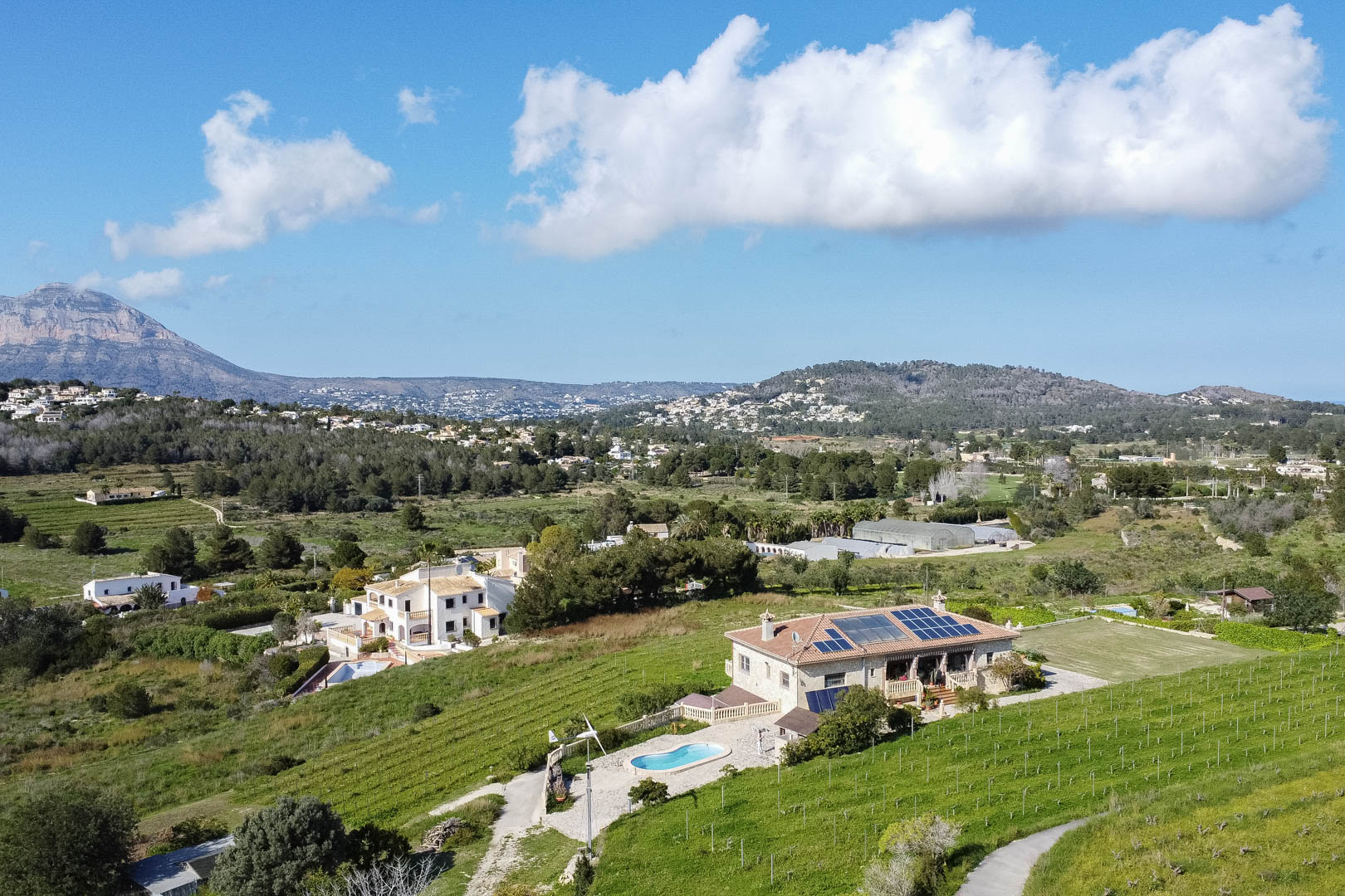 Luftaufnahme einer Steinvilla mit Solarmodulen, privatem Pool und Windkraftanlage, umgeben von Weinbergen und einer beeindruckenden Bergkulisse.