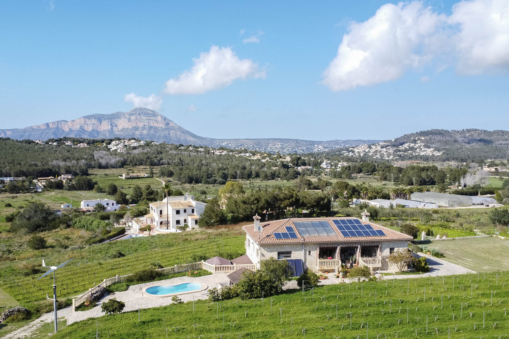 Luftaufnahme eines Finca-Anwesens mit Photovoltaikanlage, Windrad und Pool, umgeben von weitläufigen Grünflächen und einer markanten Bergkette.