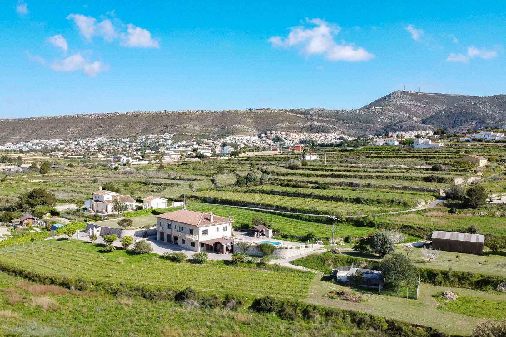 Luftaufnahme eines zweistöckigen Steinhauses mit Pool auf einem terrassierten Agrargrundstück vor einer bergigen Kulisse.