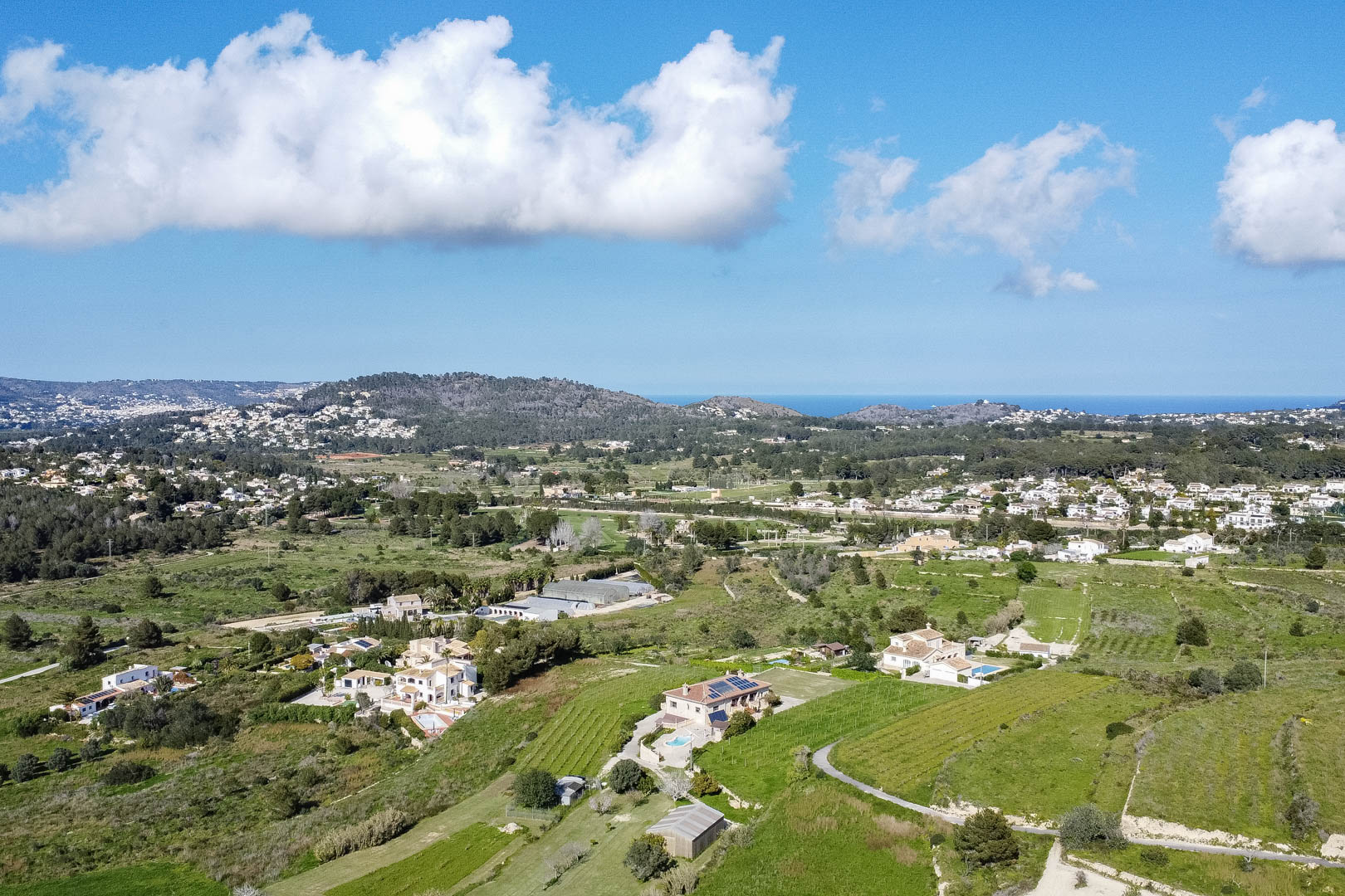Panorama-Luftaufnahme der mediterranen Landschaft mit grünen Tälern, verstreuten Villen mit Pools und weitem Blick auf das Meer am Horizont.