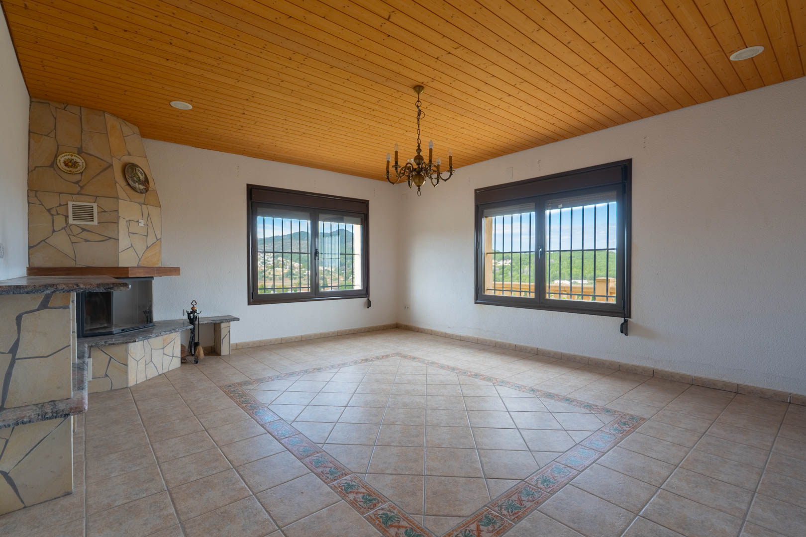 Wohnzimmer mit Holzdecke, gemauertem Eckkamin und Fliesenboden mit dekorativem Mosaik. Die Fenster bieten einen weiten Blick auf die umliegende Hügellandschaft.