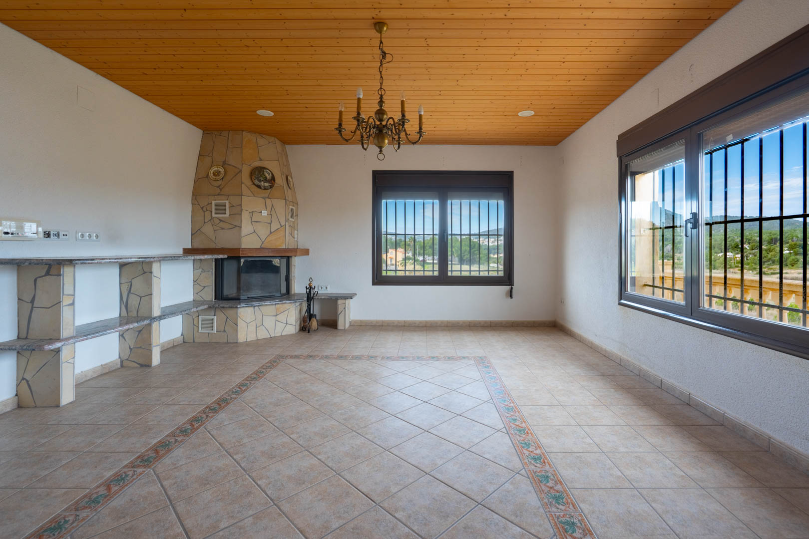 Wohnzimmer mit Holzdecke, Naturstein-Kamin und Fliesenboden mit dekorativer Bordüre. Die Fenster bieten einen weiten Blick auf die Landschaft.