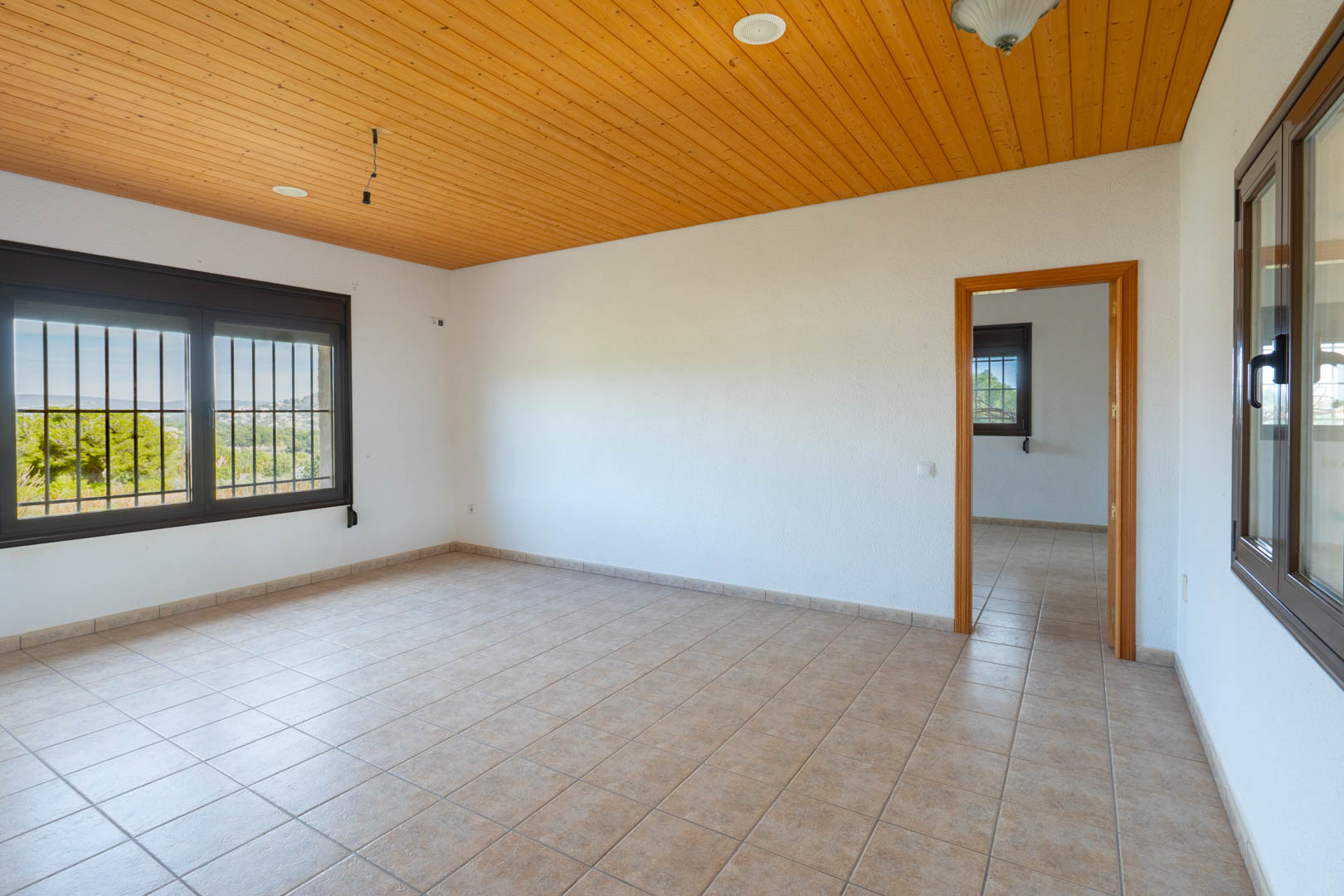 Zimmer mit Holzvertäfelung an der Decke, beigen Fliesenböden und dunkel gerahmten Fenstern mit Sicherheitsgittern und Blick in die Natur.