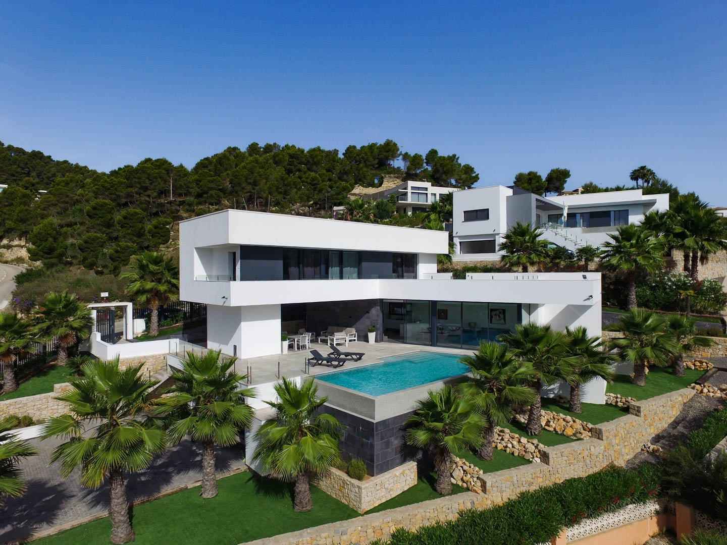 Außenansicht der Villa mit weißer kubischer Architektur, integriertem Infinity-Pool und mediterranem Garten mit Palmen und Natursteinmauern.