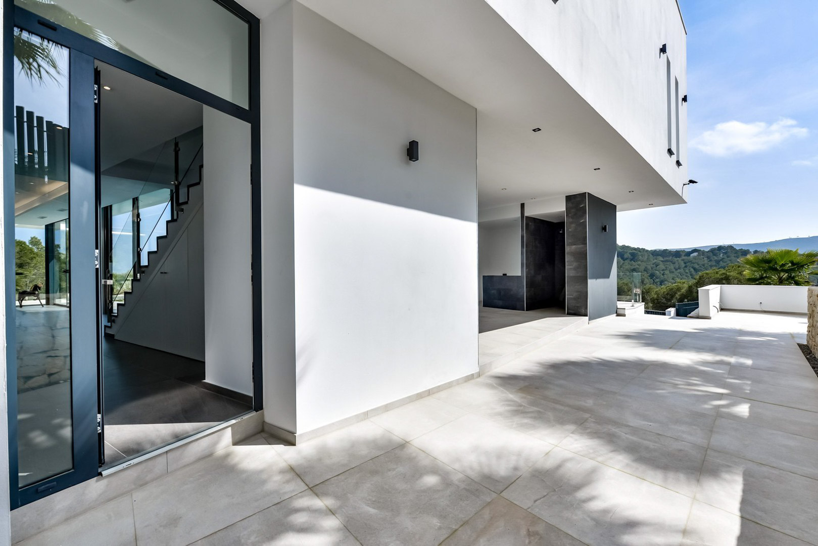 Haupteingang und überdachte Terrasse mit minimalistischer weißer Architektur, Feinsteinzeugböden und Panoramablick auf die umliegenden Hügel.