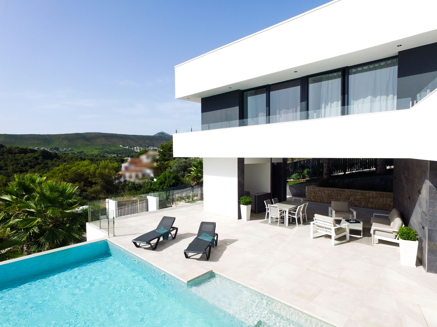Hauptterrasse mit Infinity-Pool, Sonnenliegen und überdachtem Loungebereich unter dem auskragenden Obergeschoss mit Blick auf die umliegenden Hügel.