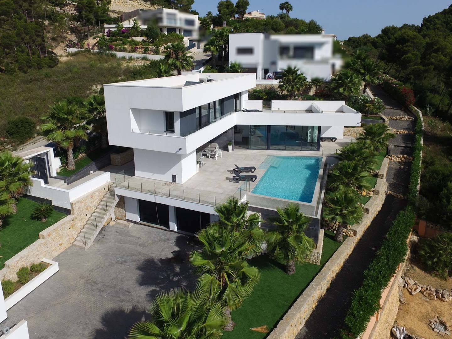Luftaufnahme der modernen Villa mit weißer Architektur, Infinity-Pool, Steinterrassen und mediterranem Garten mit Palmen.