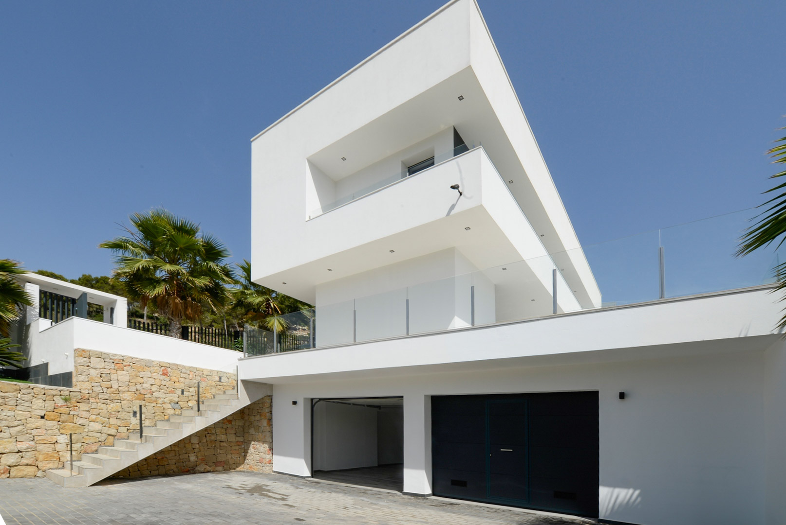 Moderne Außenansicht mit weißen kubischen Formen, Doppelgarage, Natursteinmauern und Außentreppe mit Glasgeländer.