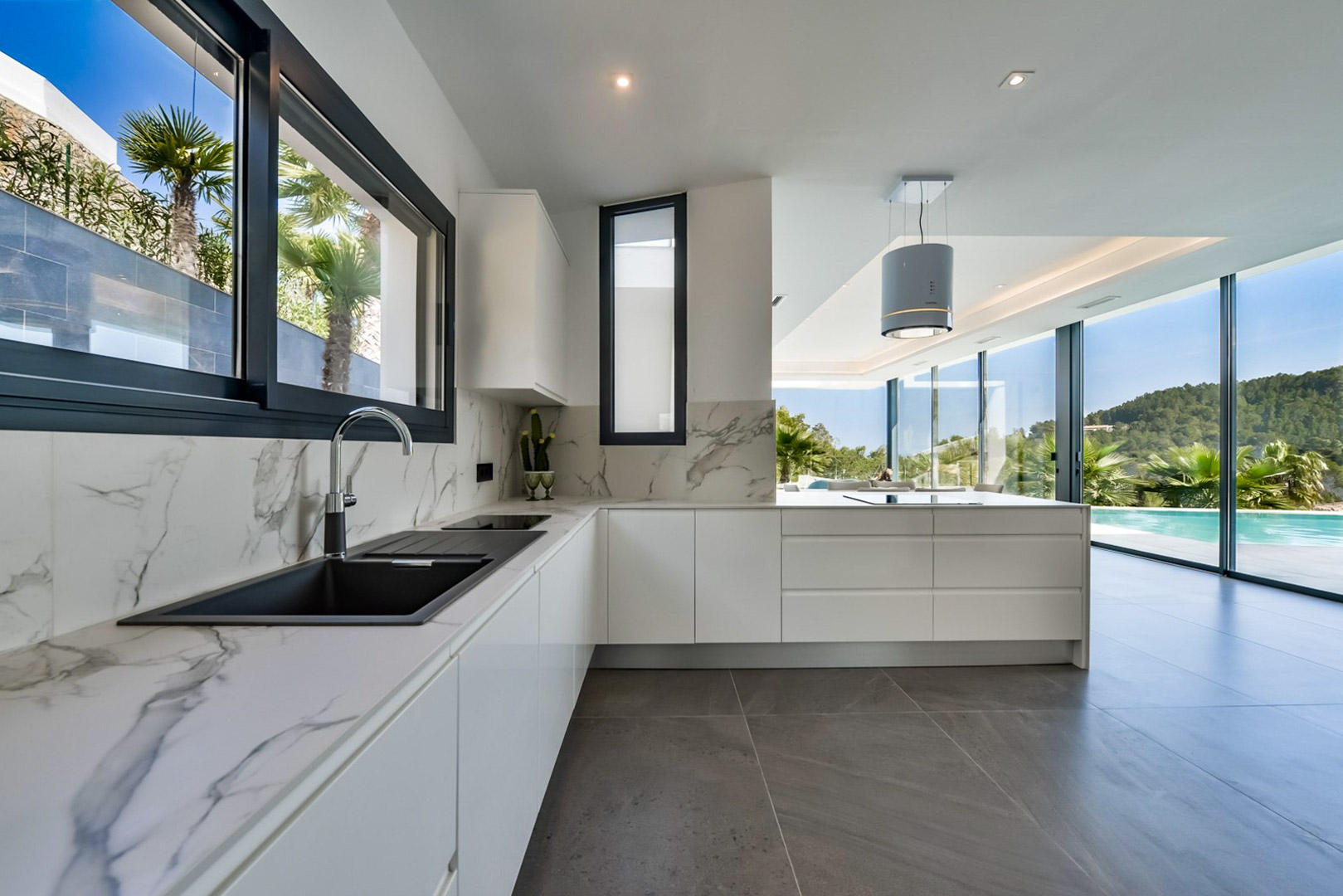 Moderne Küche mit Arbeitsplatten in Marmoroptik und zylindrischer Dunstabzugshaube. Bodentiefe Fensterfronten verbinden den Raum mit dem Außenbereich und dem Pool.